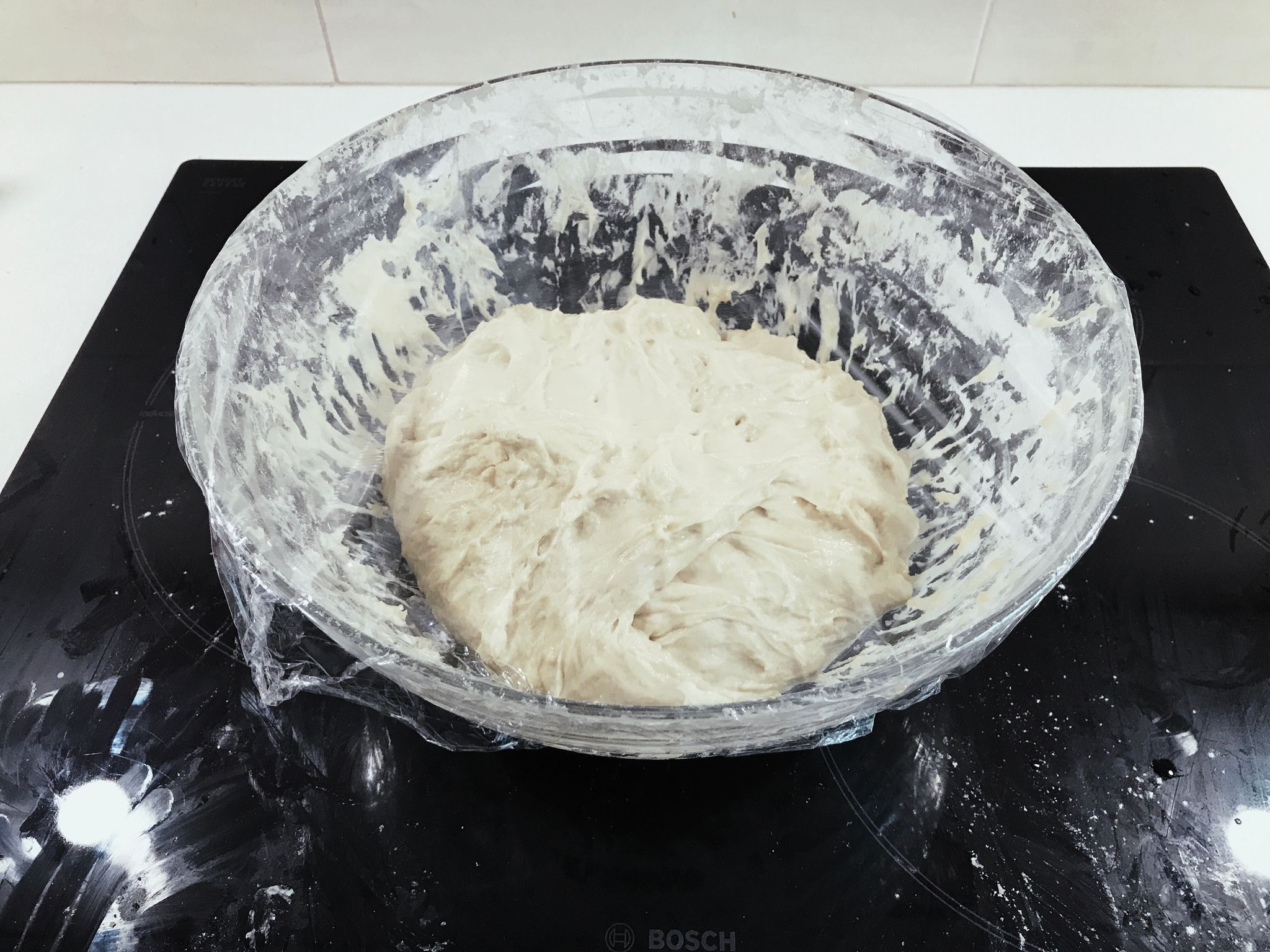 A glass bowl with a full bread mix of flour, water, salt, and yeast.