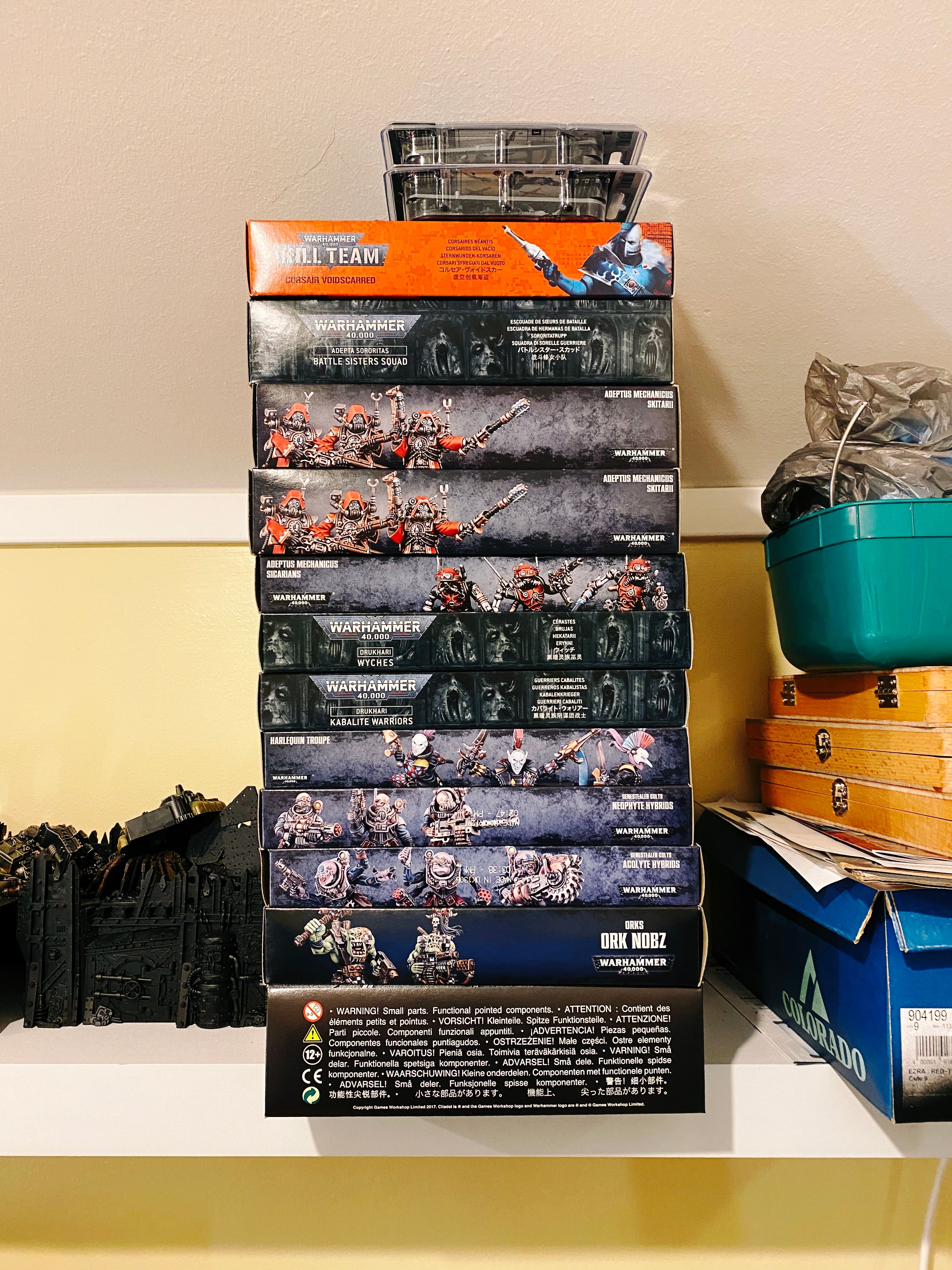 A photo of twelve boxes of unopened Warhammer 40,000 squads sitting on a shelf (plus two individual miniatures on top of that).