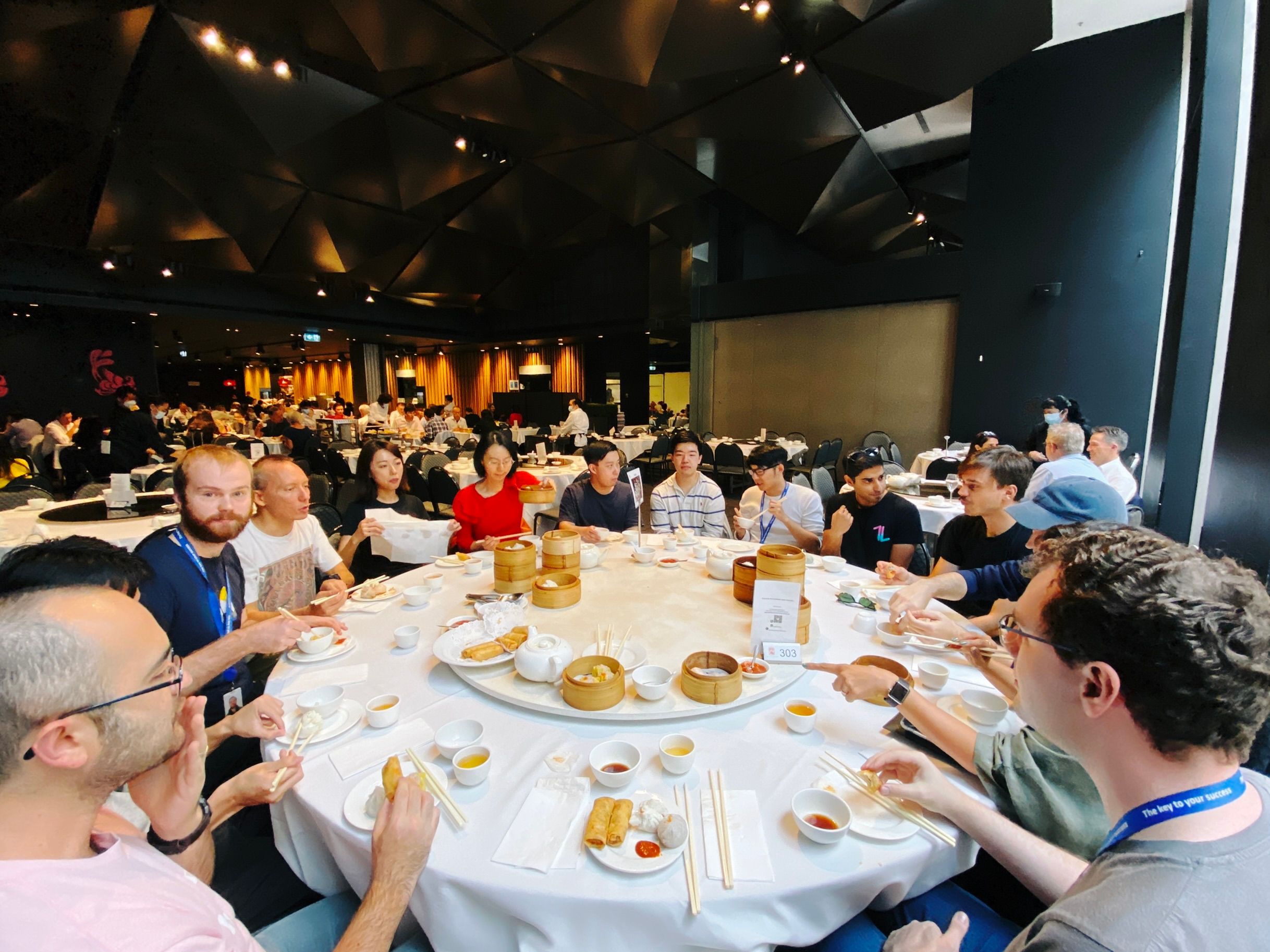 A wide-angle photo taken of a big round table with people sitting all around it and lots of food in the middle. I think yum cha is called dim sum in the US?
