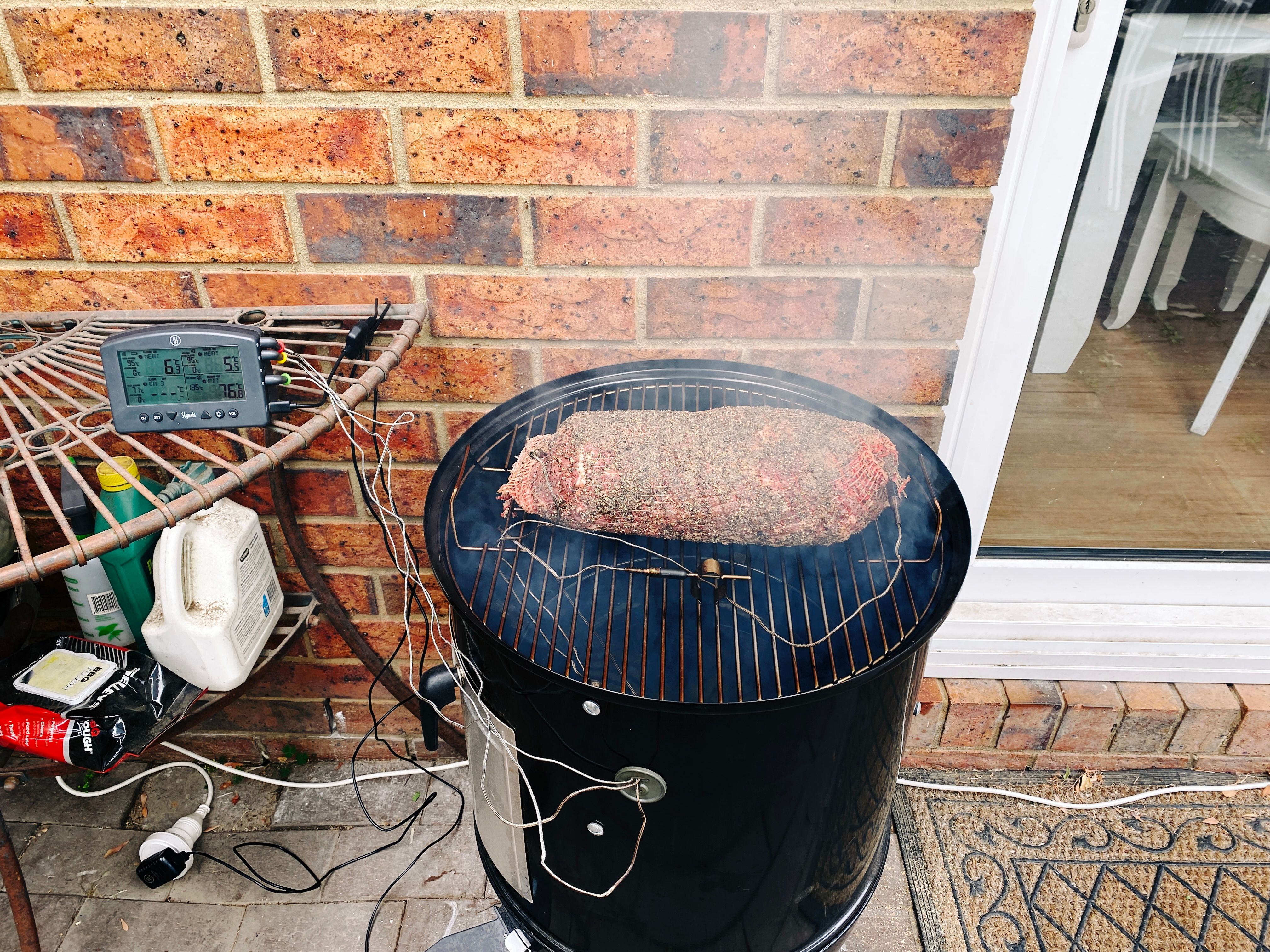 A photo of a raw 3kg pork collar butt wrapped in netting and covered in coarse pepper and salt, sitting on the grille of a smoker. There's two temperature probes going into the meat and a third measuring the ambient temperature of the smoker itself, all three of which are going into a little box with an LCD (a Thermoworks "Signals") that shows the temperature of each probe. The wood has started smoking already and there's lots of it coming up from underneath the grille.