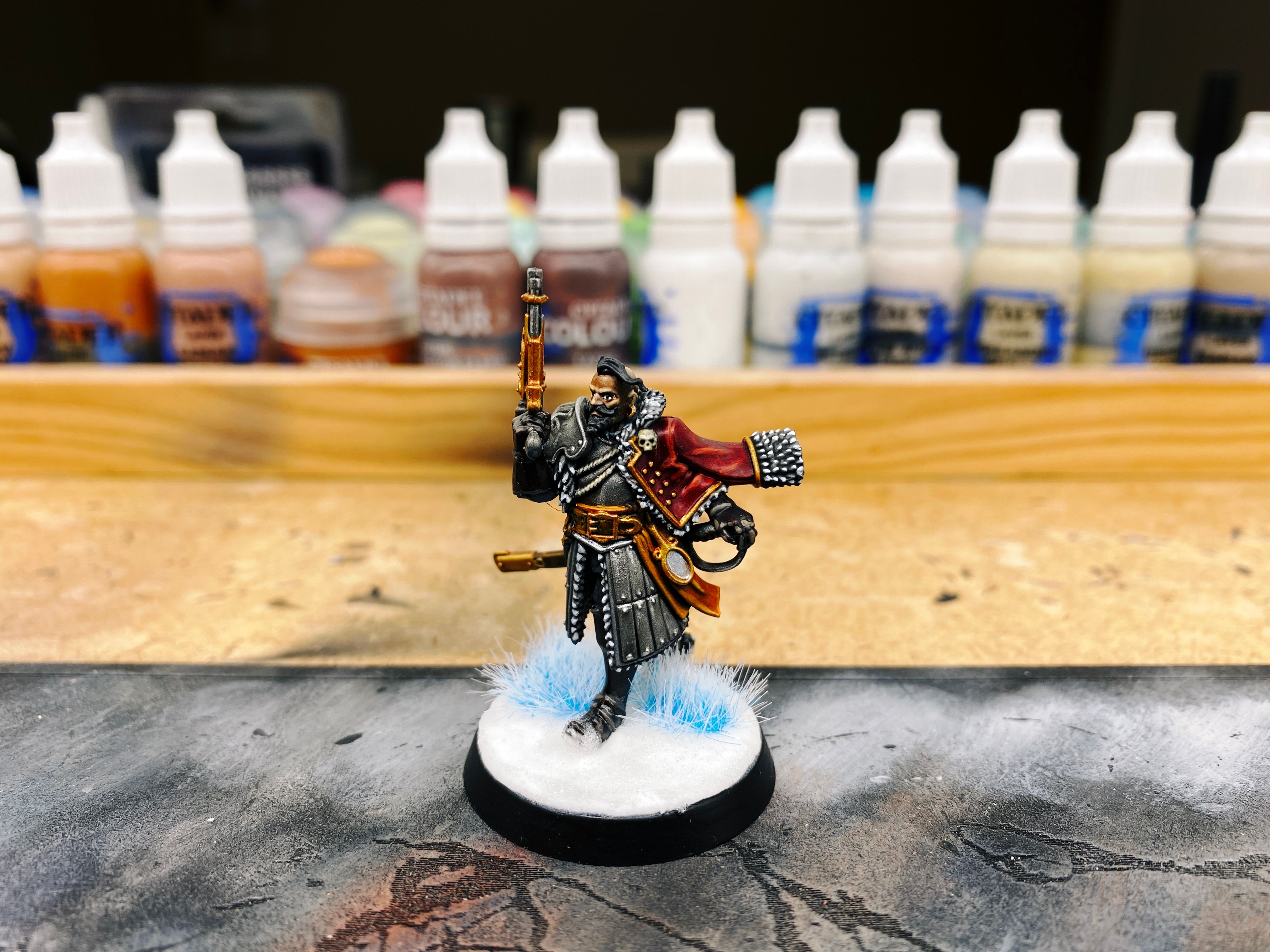 A photo of an aristocratic-looking miniature mid-stride. He's got a magnificent beard and mustache that's curled up at the ends, plus a long top-knot of hair and the rest of his head is bald. He's holding a steampunk-looking pistol upright and has a red jacket edged with white fur billowing out behind him, and his other hand is on the pommel of his sword. His robe thing is lined with armour plates and he's wearing an armoured chestpiece as well.