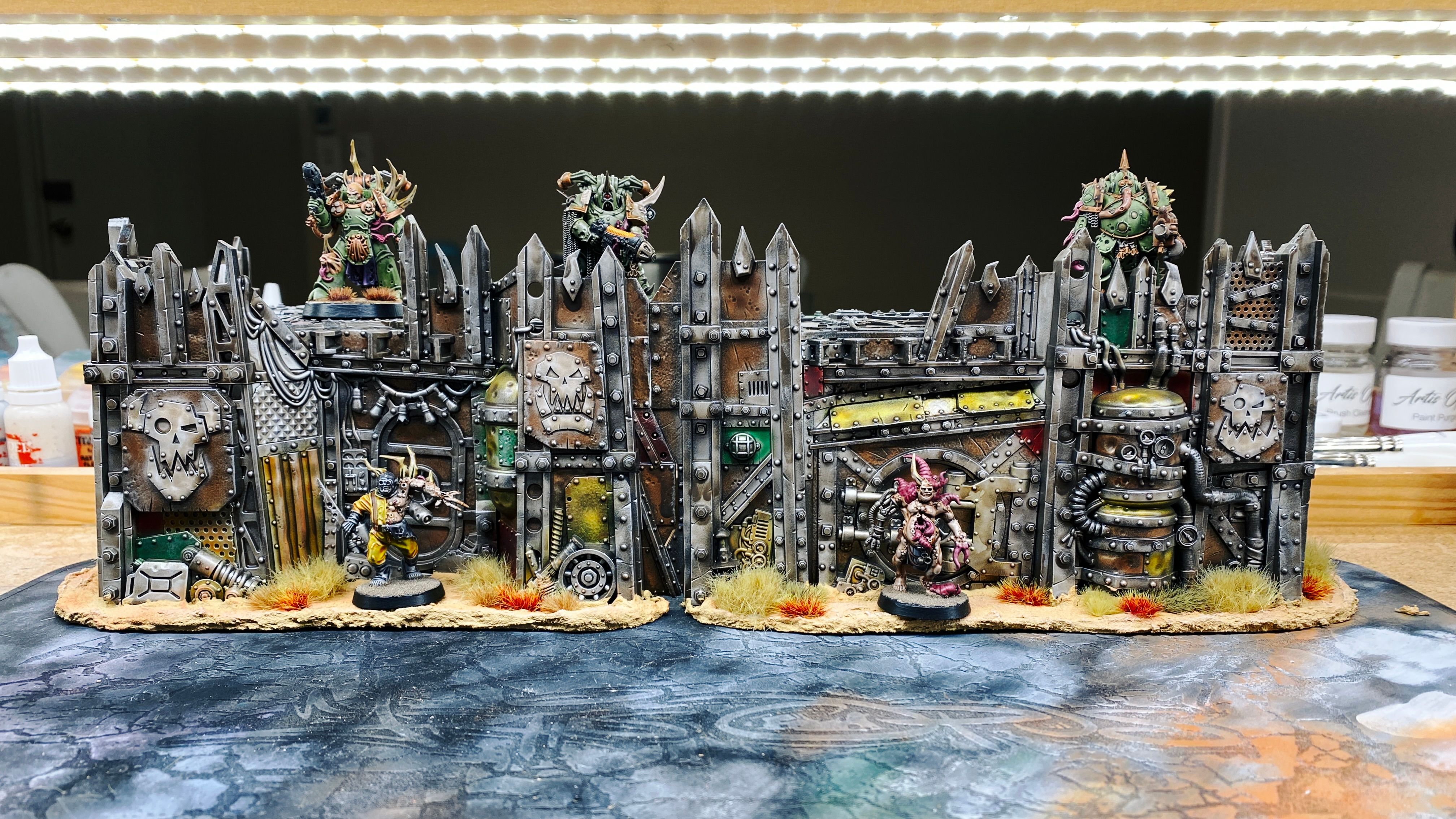 A photo of two of the EXTREMELY ramshackle pieces of terrain from the Warhammer 40,000 Kill Team: Octarius box from Games Workshop. They're parts of an Ork fort and look like they belong in a junk yard, the walls and platform on top are made up of a whole ton of pieces of metal scrap, like big plates, corrugated pieces, massive beams, there's a door in there, and a couple of empty gas cylinders. Both pieces are on cut-out pieces of cardboard that are covered in a texture paint that makes it look like sand, and there's various tufts of dry grasses growing around the base of the walls.