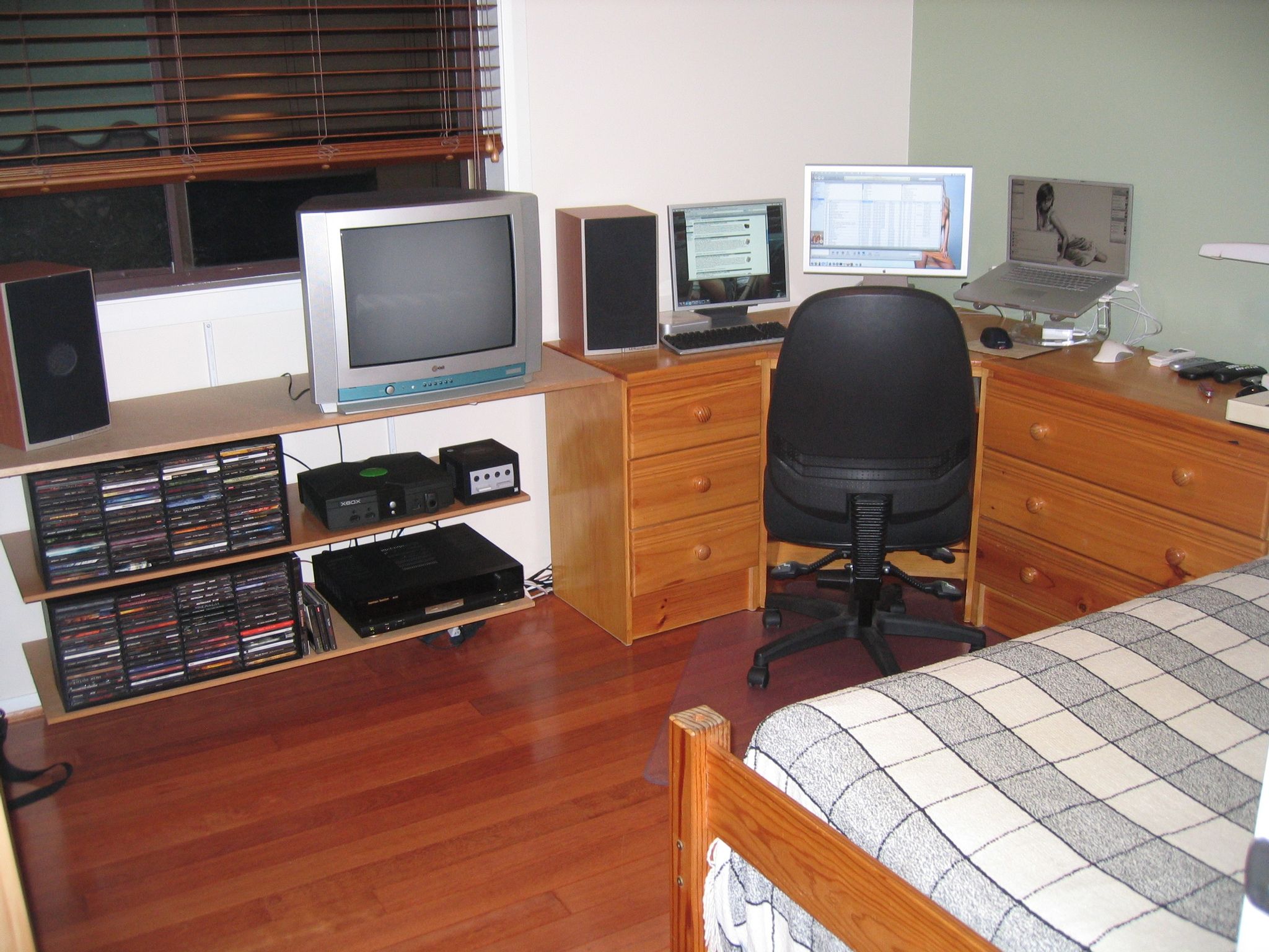 A photo of my room. The floor is reddish wood, there's a corner desk towards the right with a 20" Apple Cinema Display in the middle, a 17" PowerBook G4 to the right, and a 17" 4:3 monitor the left. To the left of the desk are three wall-mounted shelves with a CRT TV on the top, an Xbox and GameCube plus a rack of CDs in the middle, and another rack of CDs on the bottom alongside a stereo amplifier.