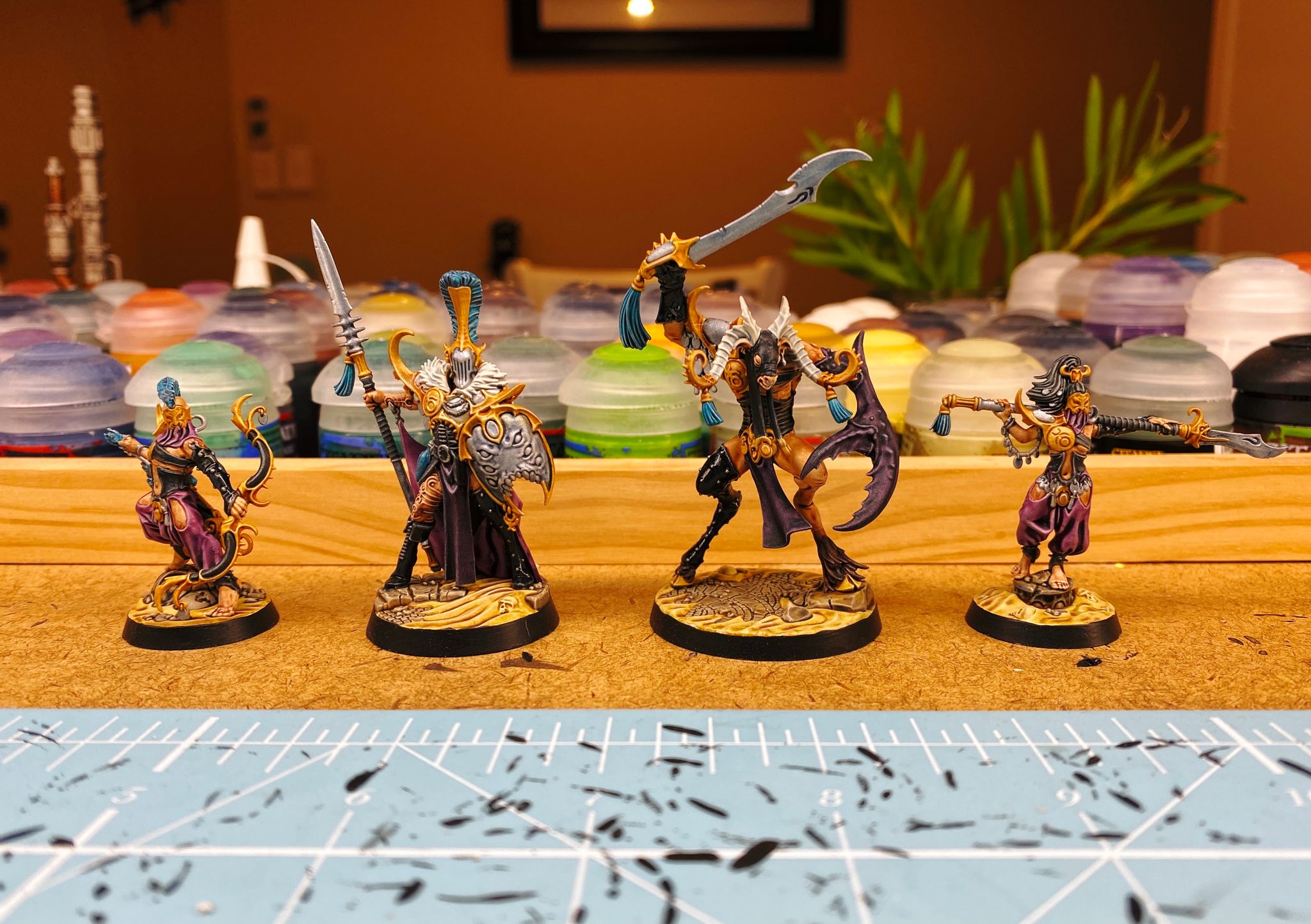 A photo of the 4 miniatures of the Dread Pageant from Warhammer Underworlds: Direchasm. 2 are human, male and female, both in puffy purple Middle-Eastern type pants, black corsets split down the front, and with gold masks on. He has an ornate bow, she is holding a glaive over her shoulders. The 3rd looks human but much taller, with a studded PVC-clad leg and holding a spear and shield with 3 sets of eyes. Last is is a beastman with a massive claw for an arm and wielding a sword, he also has a PVC-clad leg.