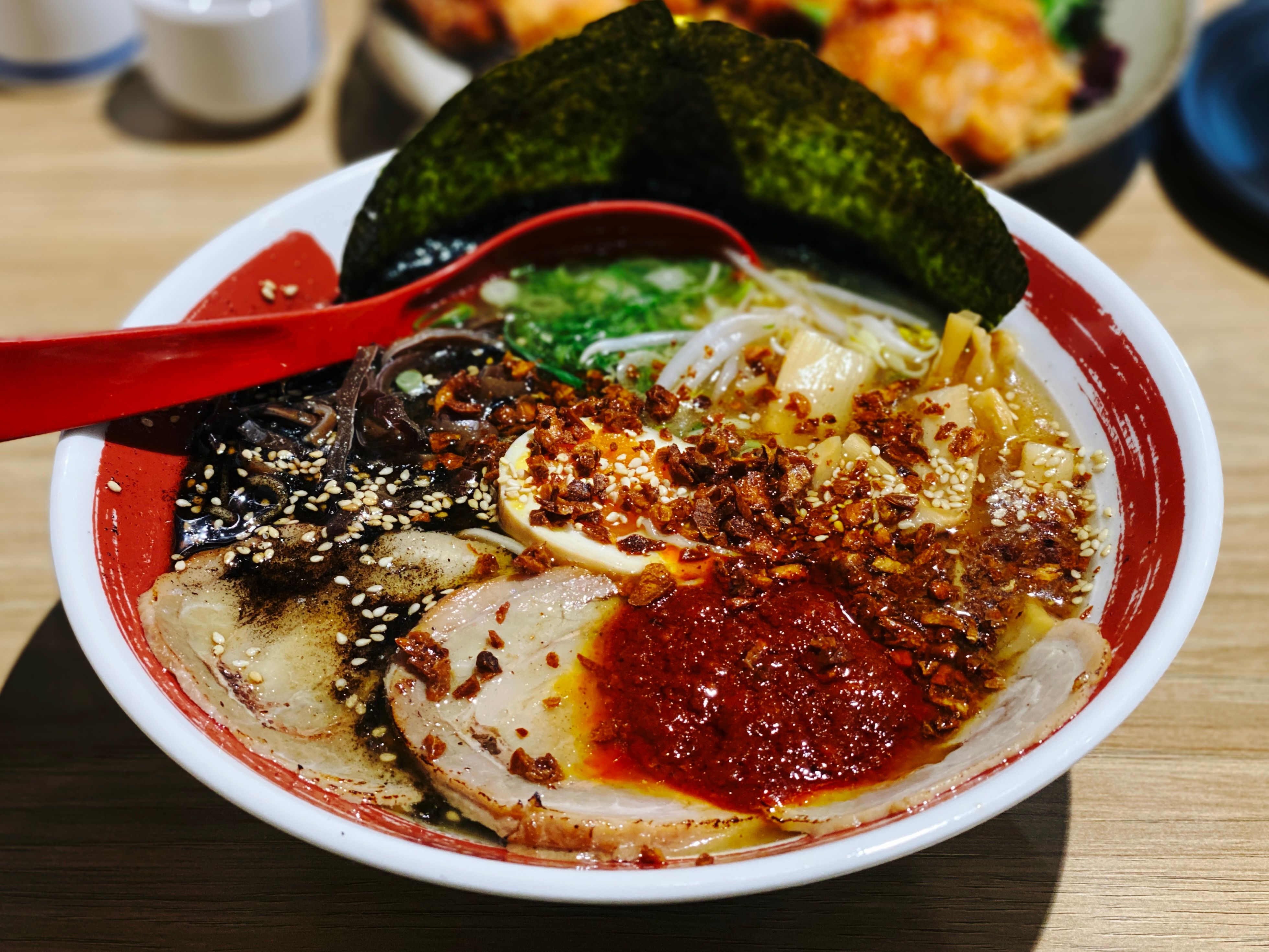 A photo of a bowl of ramen. It has slices of pork with chilli sauce on it, shallots, some sort of thinly-sliced mushroom thing, and two pieces of nori at the back, plus the standard ramen noodles.