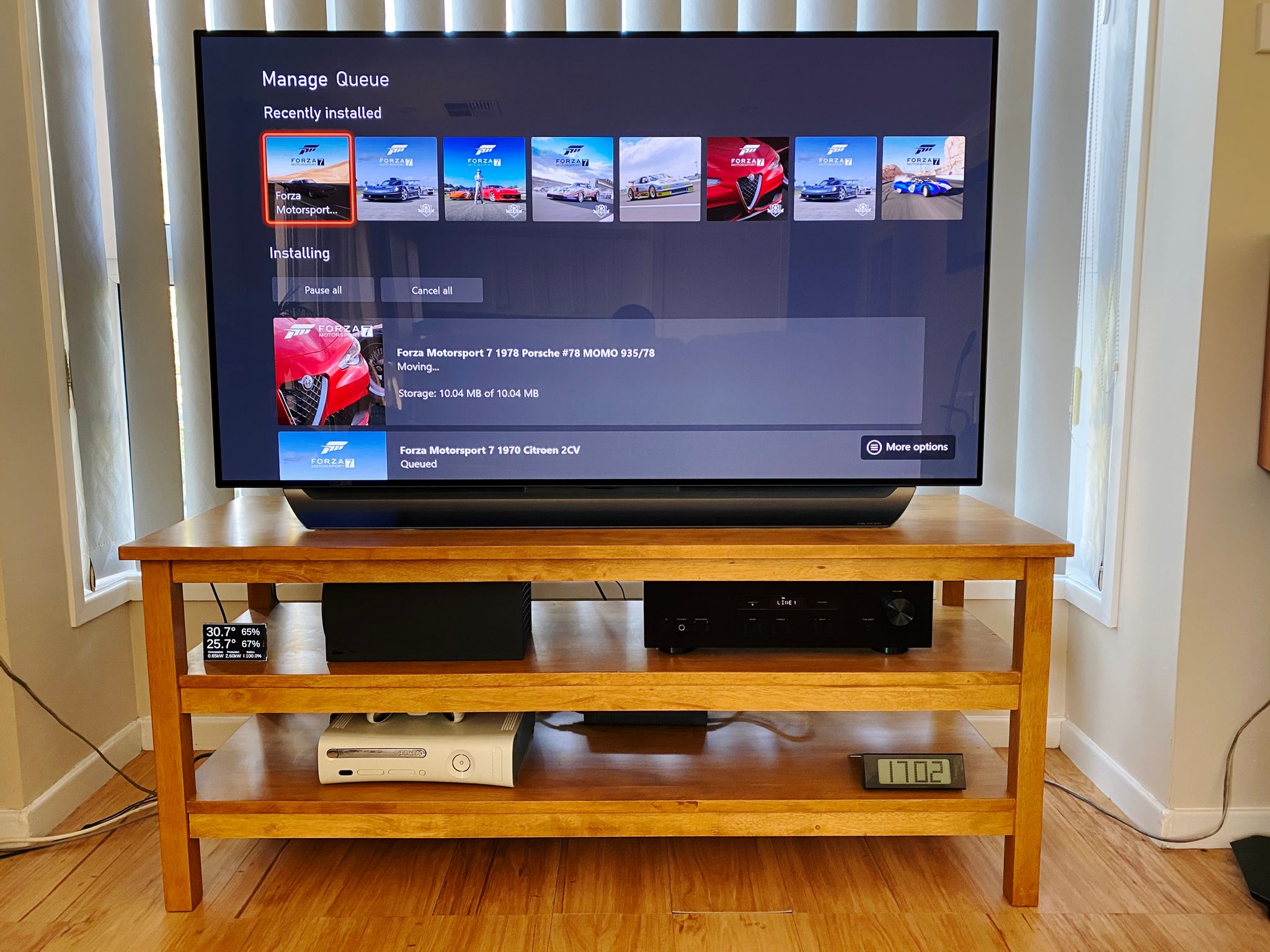 A photo of our TV setup. The TV stand is a two-shelf wooden one, the TV is sitting on top, on the shelf underneath is the Xbox Series X sitting on its side next to the stereo amplifier, and on the bottom shelf is an Xbox 360.