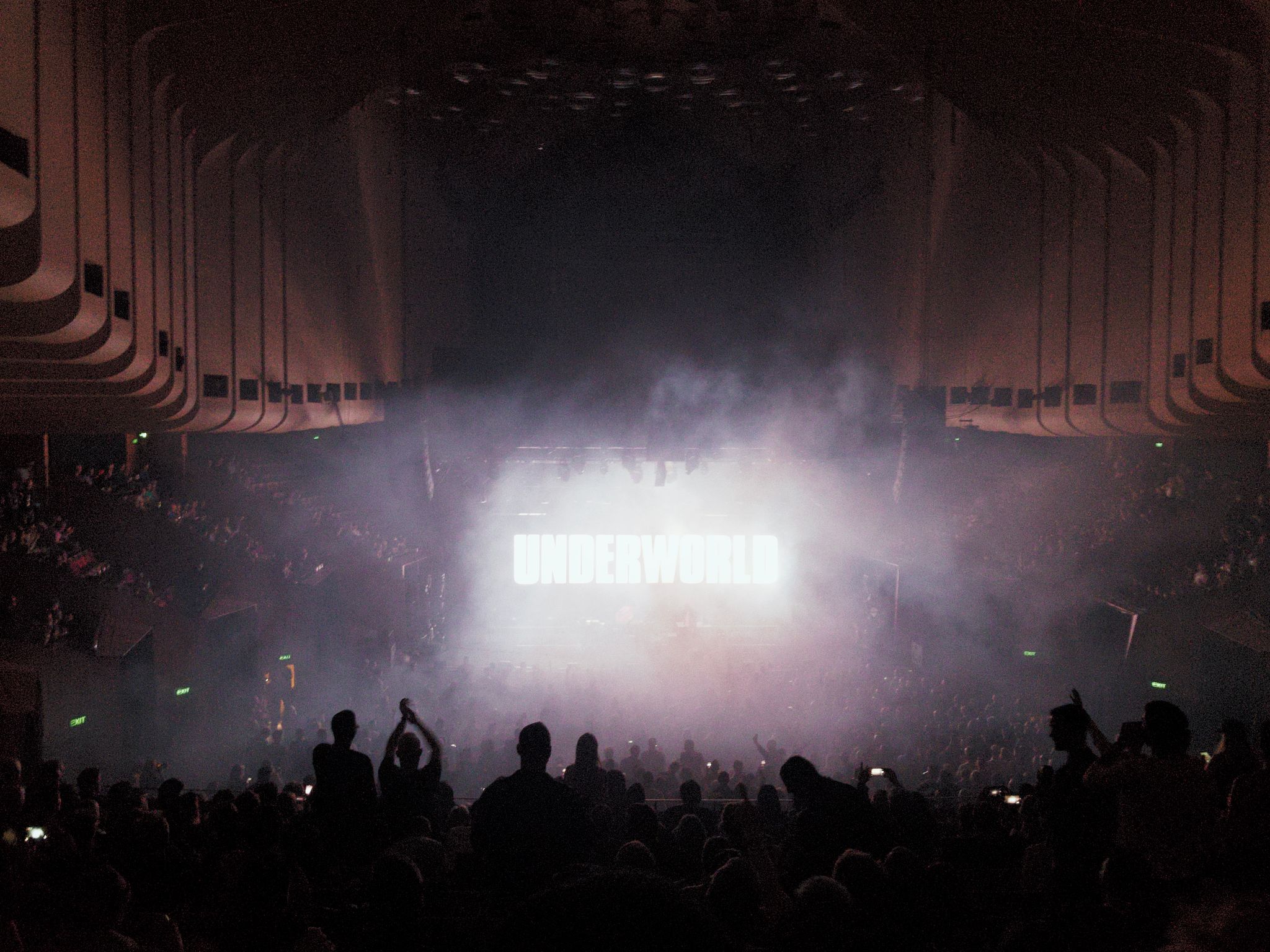 Looking down from seating inside the Opera House towards the stage, everything is hazy from smoke machines and the back of the stage has "UNDERWORLD" projected onto it in giant white letters. Silhouettes of the crowd cheering can be seen in the smoke.