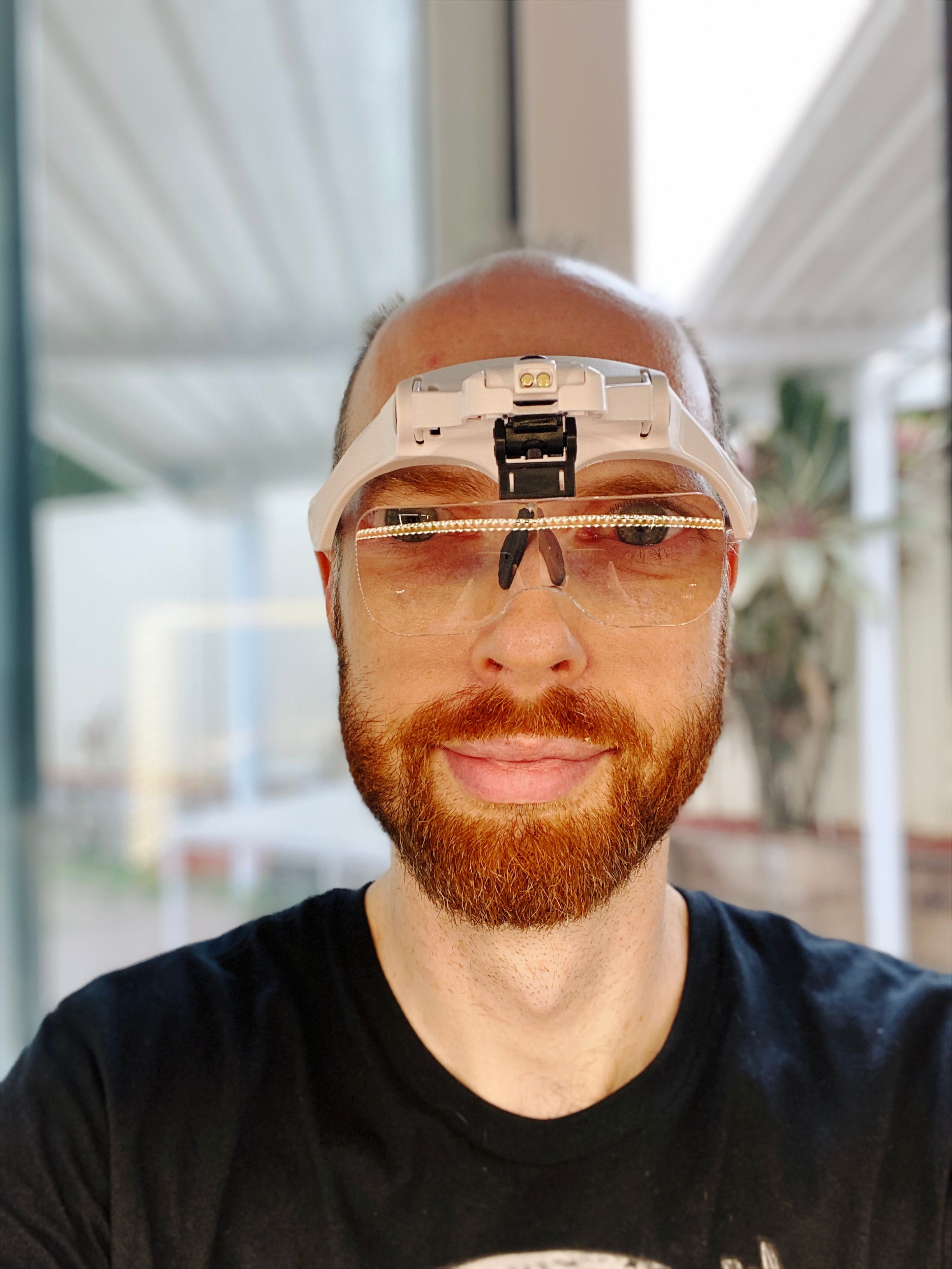 A selfie of me, a white man with a short red beard and receeding hairline, wearing head-mounted set of magnifying eyeglasses. The headset itself is white and the lenses are a single piece of almost sunglasses-shaped hard plastic that attaches to the front of the headset and can be moved forwards or backwards. My eyes look freakishly large because I'm looking directly through the lenses at the camera.