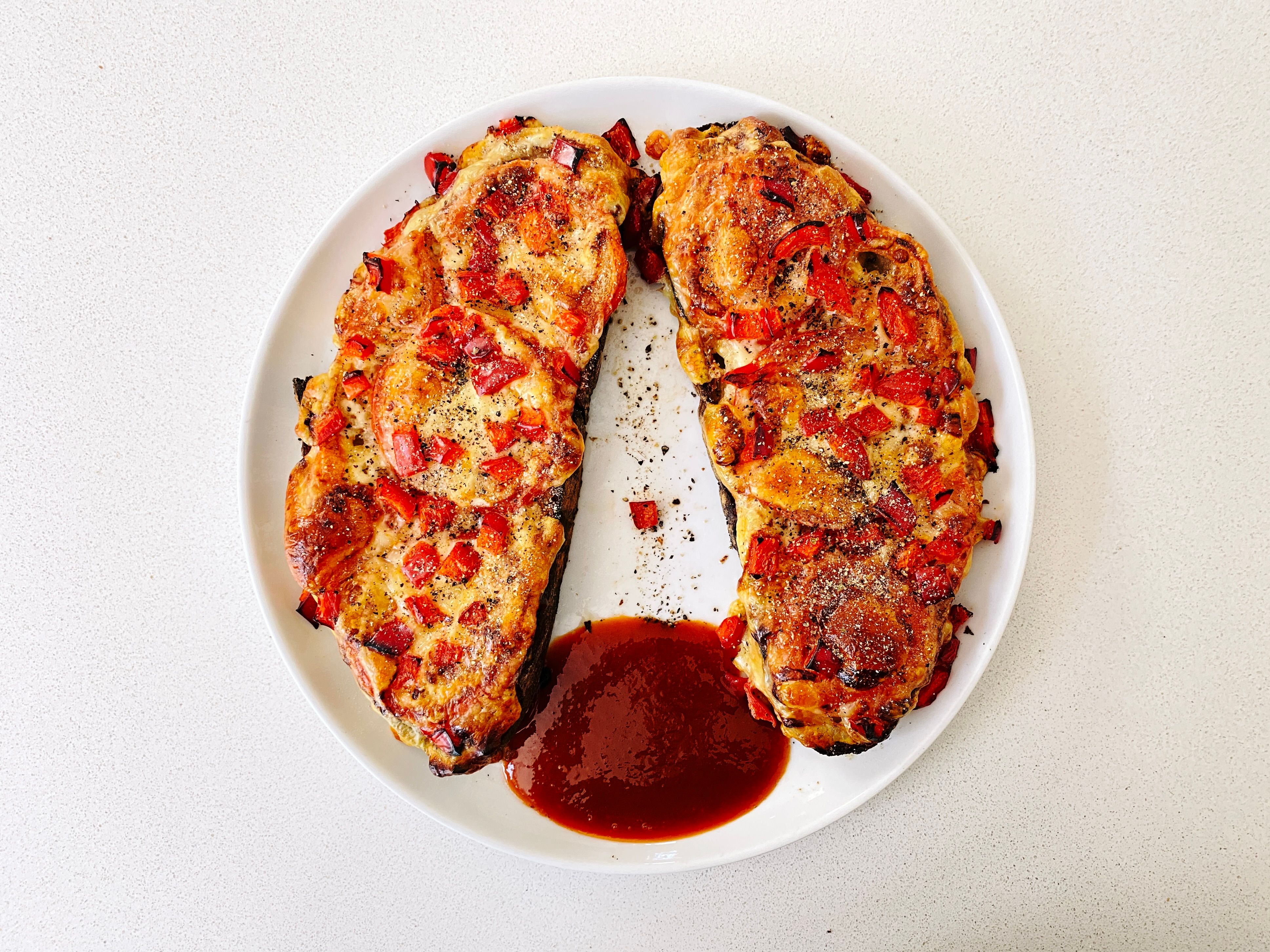 A photo of two slices of toasted bread with tomato and golden-brown cheese and loads of capsicum on top, covered in pepper, sitting on a plate. There's a big squirt of Tabasco-brand sriracha sauce on the plate in between the two slices of bread.