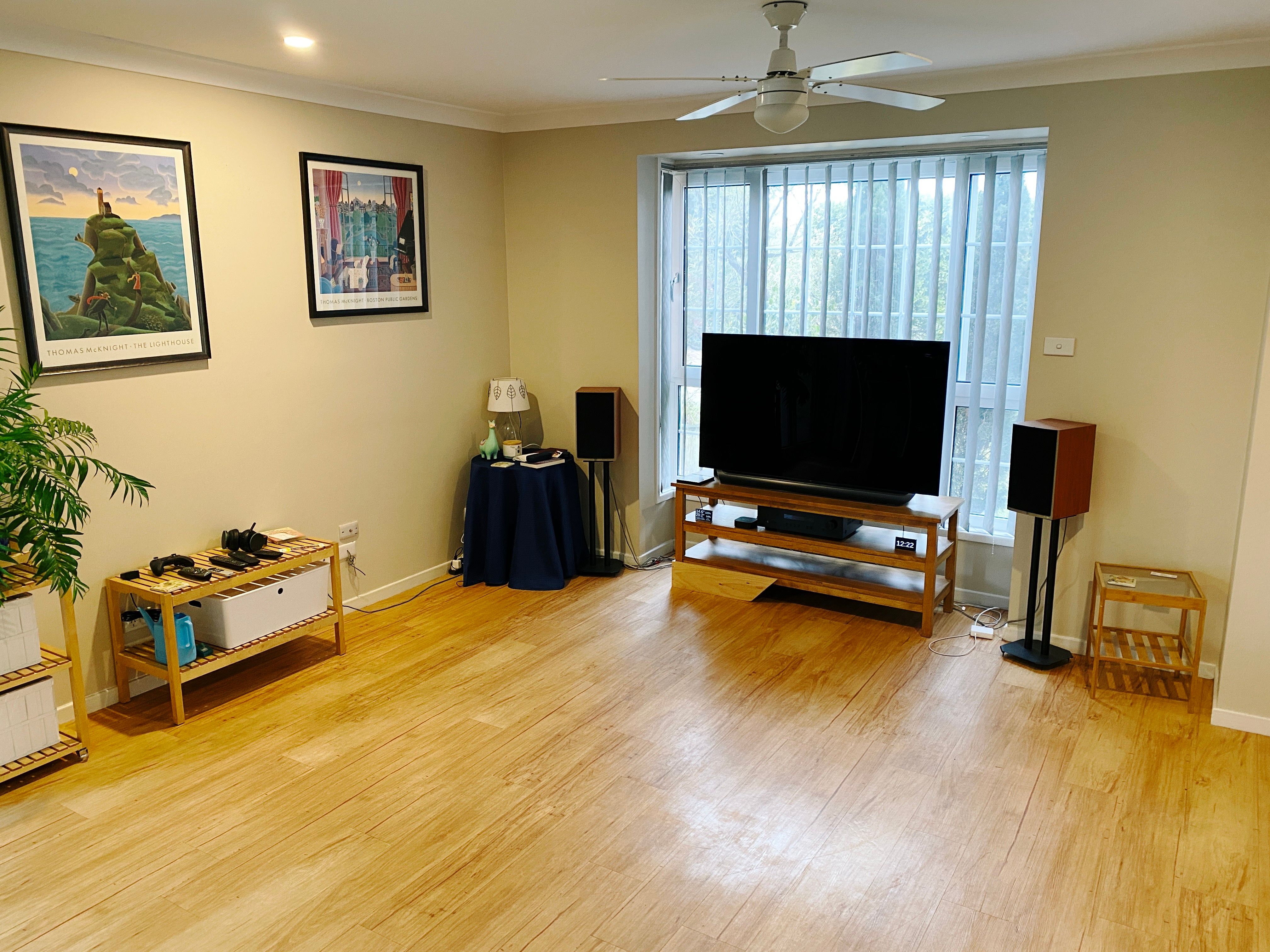 A photo of our lounge room. There's a box window towards the middle of the photo with a wooden TV stand and TV in front of it and bookshelf speakers flanking the TV, a round table with a lamp and various doodads in the corner to the left of the TV stand, plus a couple of other little side tables. The middle of the room is completely empty now due to the lack of lounge.