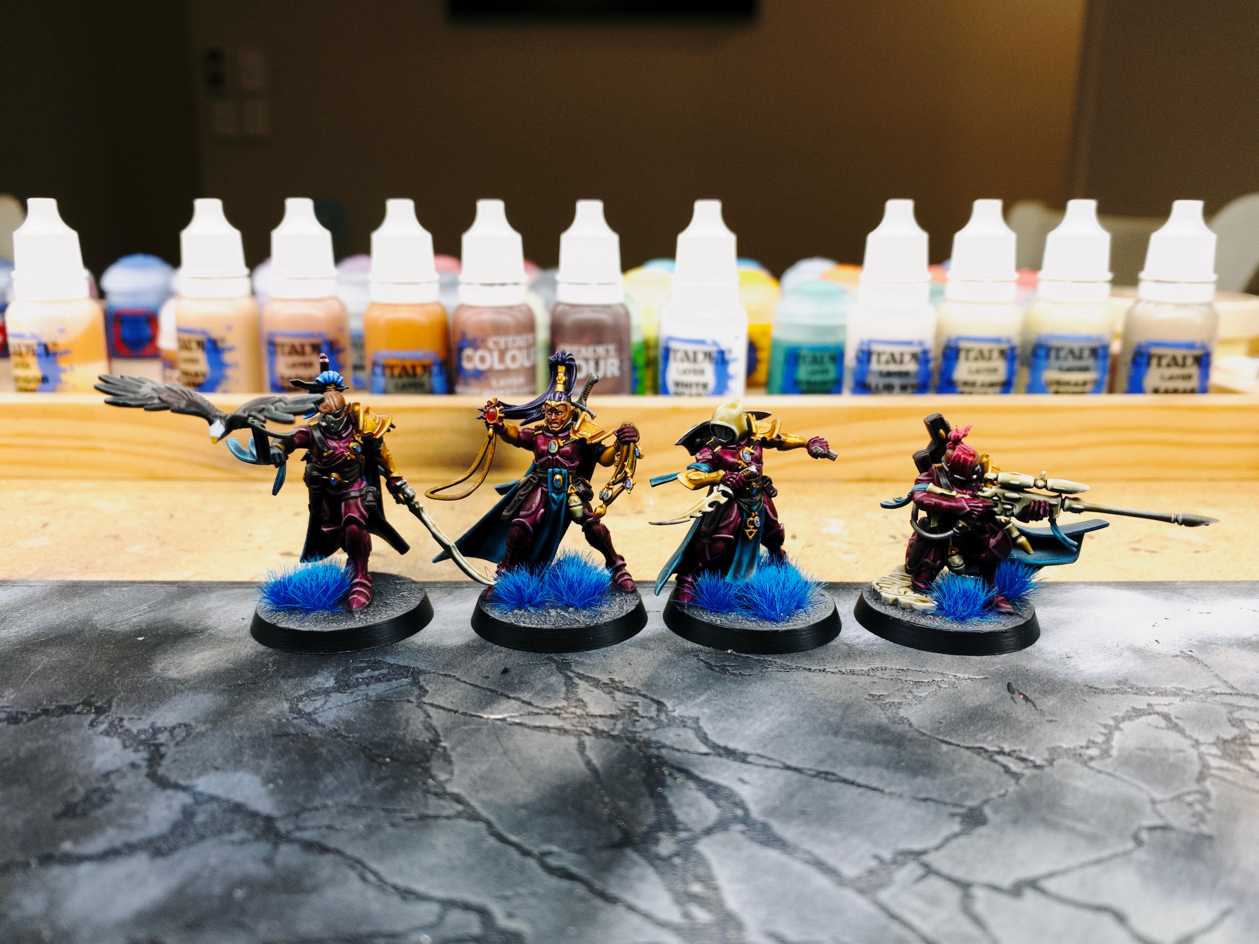 A photo of four miniatures. They're basically space elf pirates, all their armour is elegant and flowing and a rich burgundy colour, and they have cool turquoise capes and three of them have either a full mask or rebreathers on. The first the has a long sword in one hand and has a bird that's just taken flight from his other hand, the second is holding up a big gem in front of her in the same sort of way that movies depict people holding up crosses to ward off vampires, the third has a hood on and is holding two short blades out on either side like she's just finished slicing someone to ribbons, and the last is kneeing while looking down the sights of a sniper rifle. All but the hooded one have very punk-looking hair: bright-coloured with top-knots and half-shaved heads.