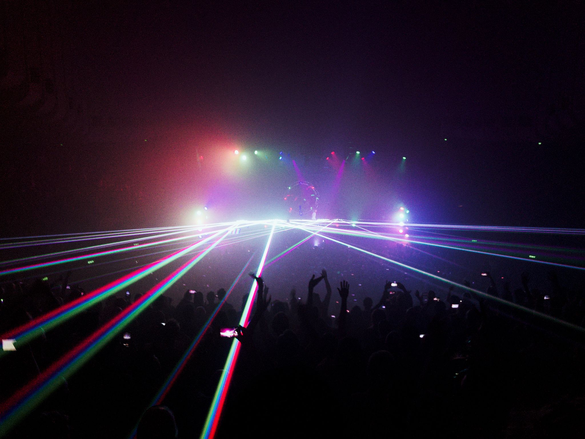 Closely-aligned lasers of red, green, and blue shooting out from the stage below the point of view of the photo. Multicoloured spotlights of the same colours and purple are shining through the smoke machine haze to make rainbow colours.
