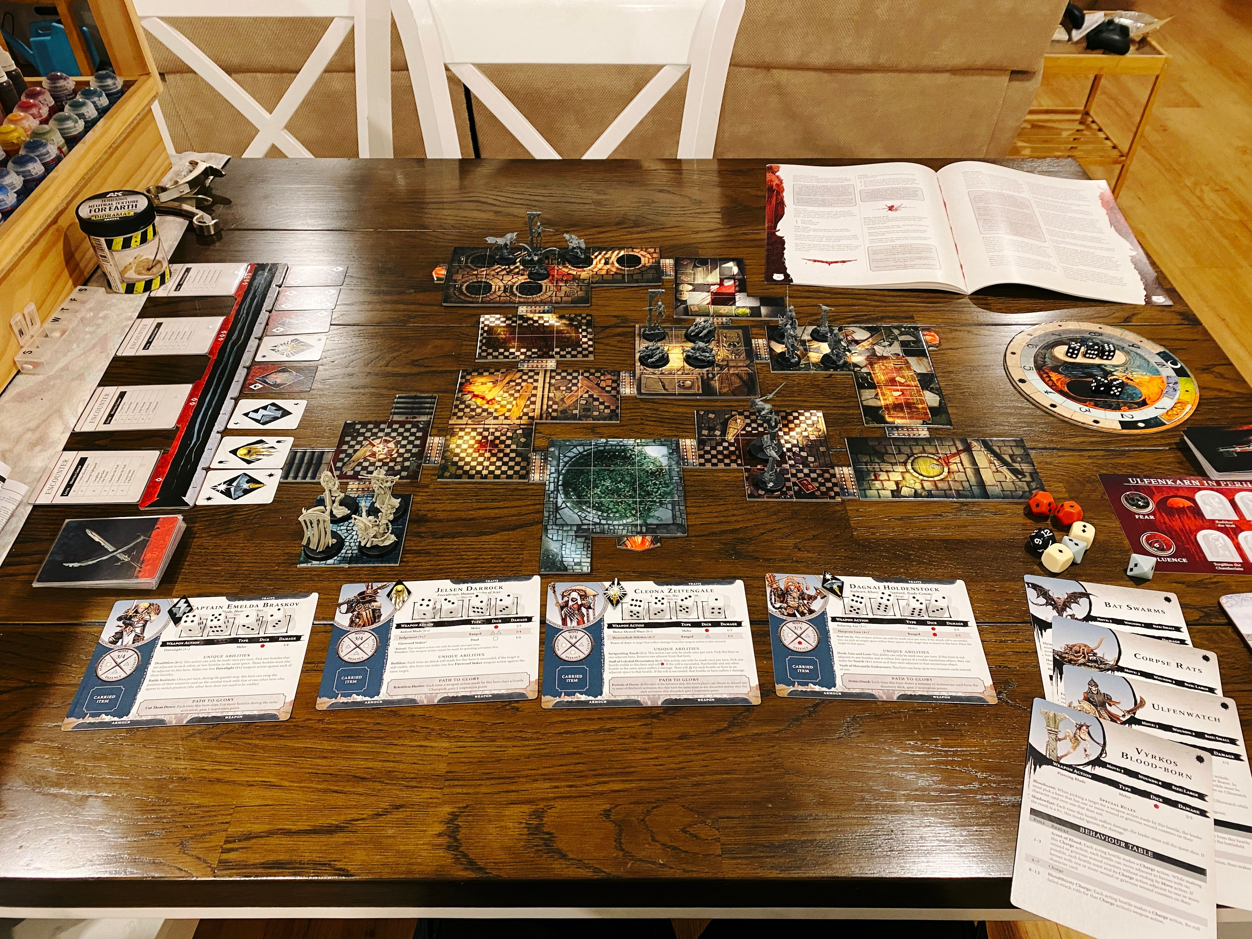 A photo of our dining table with a Warhammer Quest game set up on it. There's lots of cards and dice everywhere, and in middle are a bunch of variously-shaped tiles connected together that look like the inside of fantasy buildings. There's four hero miniatures in bone-white plastic at the bottom, and three groups of plain grey hostile miniatures at various points on the board (three skeletal warriors, three rat swarms, three bat swarms, and a lone vampire with a very long sword).
