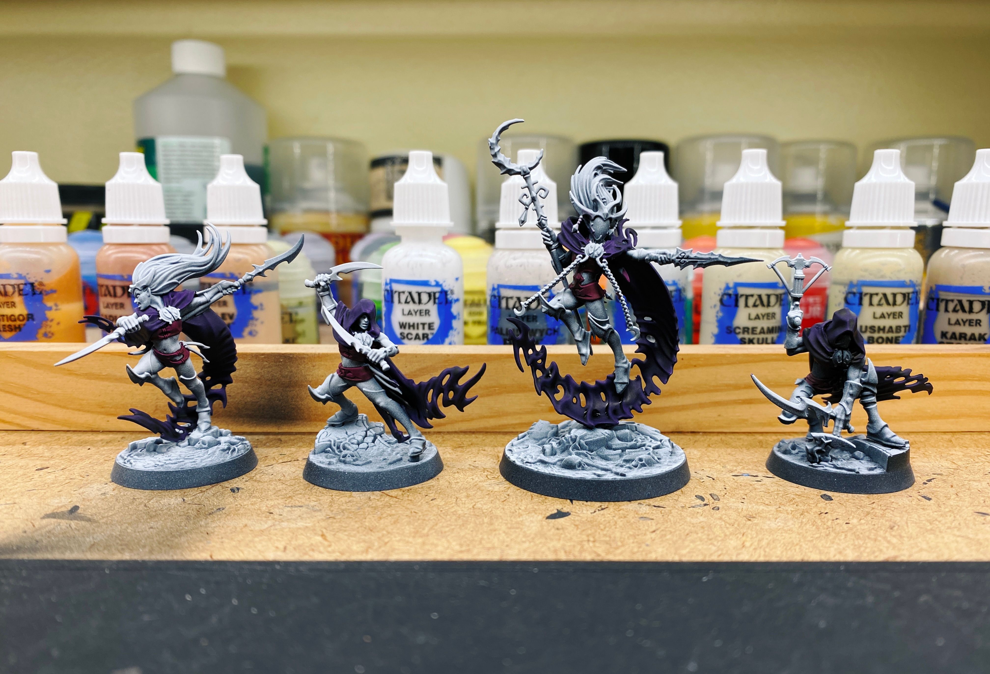 A photo of four partially painted miniatures. They're all elves in various dynamic positions and have cloaks that turn into... not quite smoke at the ends, and the cloaks have been painted this INCREDIBLY deep and rich dark purple colour.
