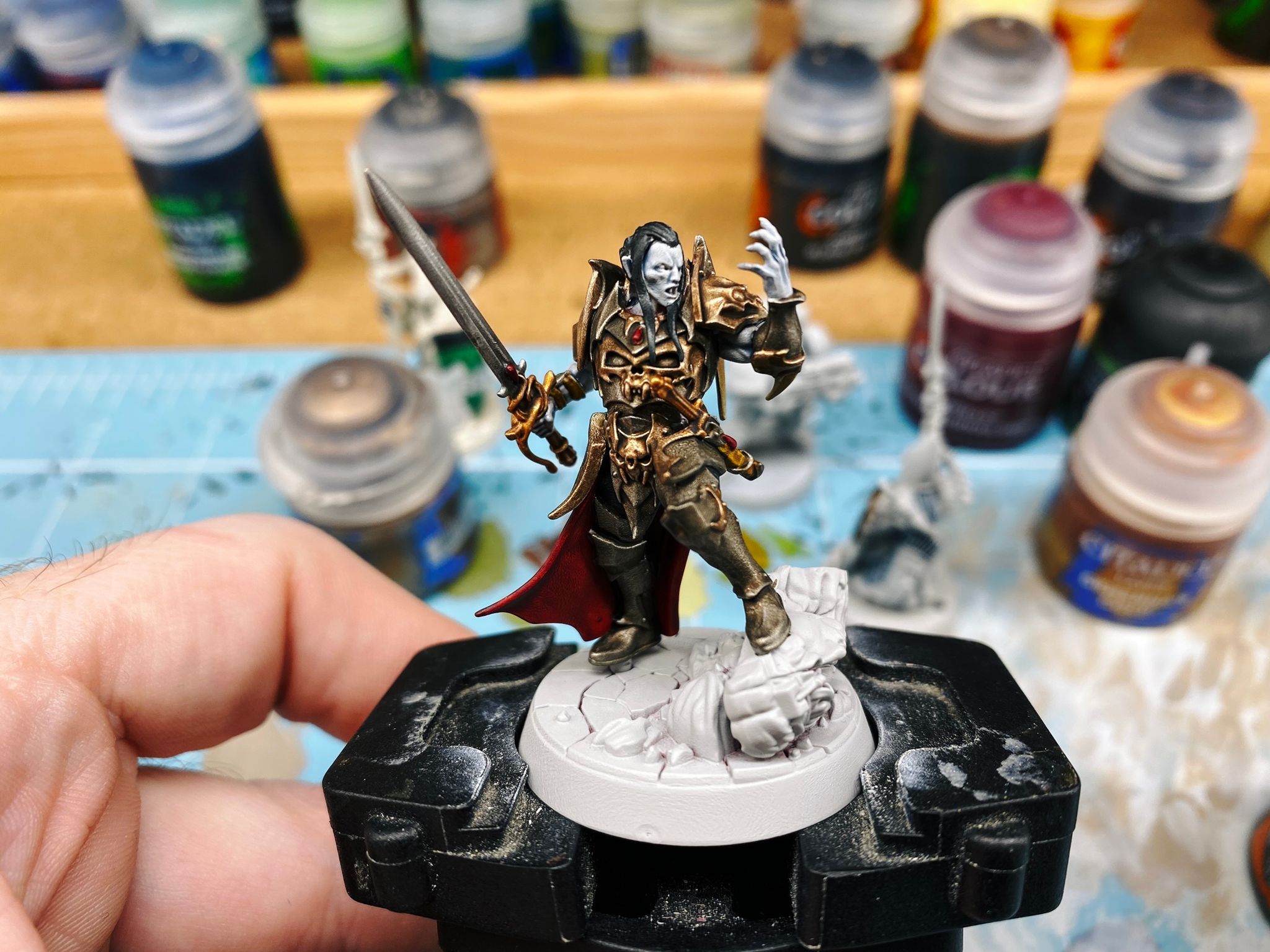 A photo of a vampire miniature, Prince Duvalle. His skin is very pale blue like he's dead (which is he, because he's a vampire), he's wearing steel- and brass-coloured armour with a blood red cloak, he has a sword in one hand, and his other hand is held up in front of him in a sort of "We will crush them" pose. His front foot is resting on top of the broken-off head of a stone statue.