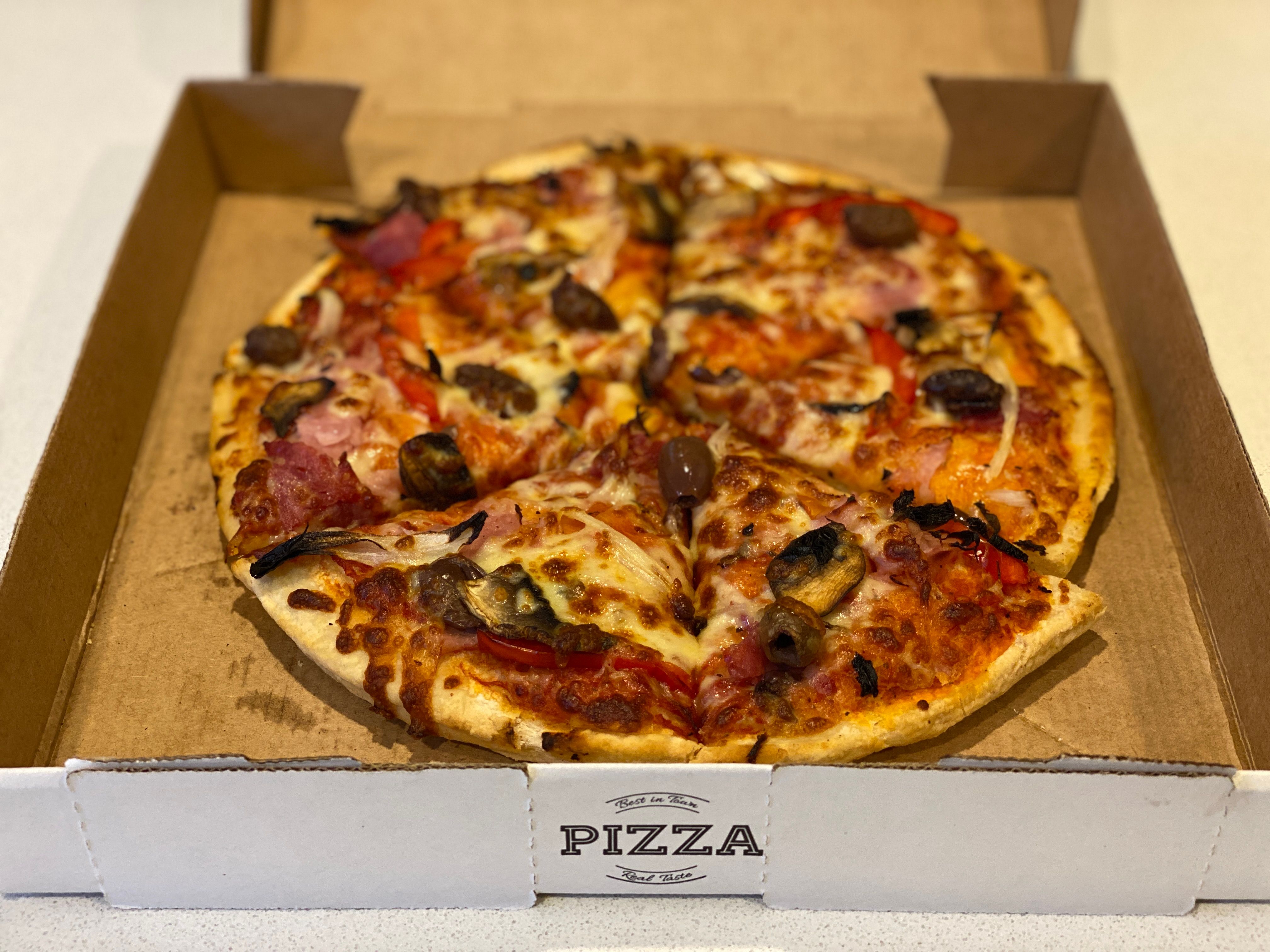 A photo of a medium-sized pizza in a cardbox takeaway pizza box. It's got ham, mushrooms, capsicum, and olives on top in addition to the standard cheese.