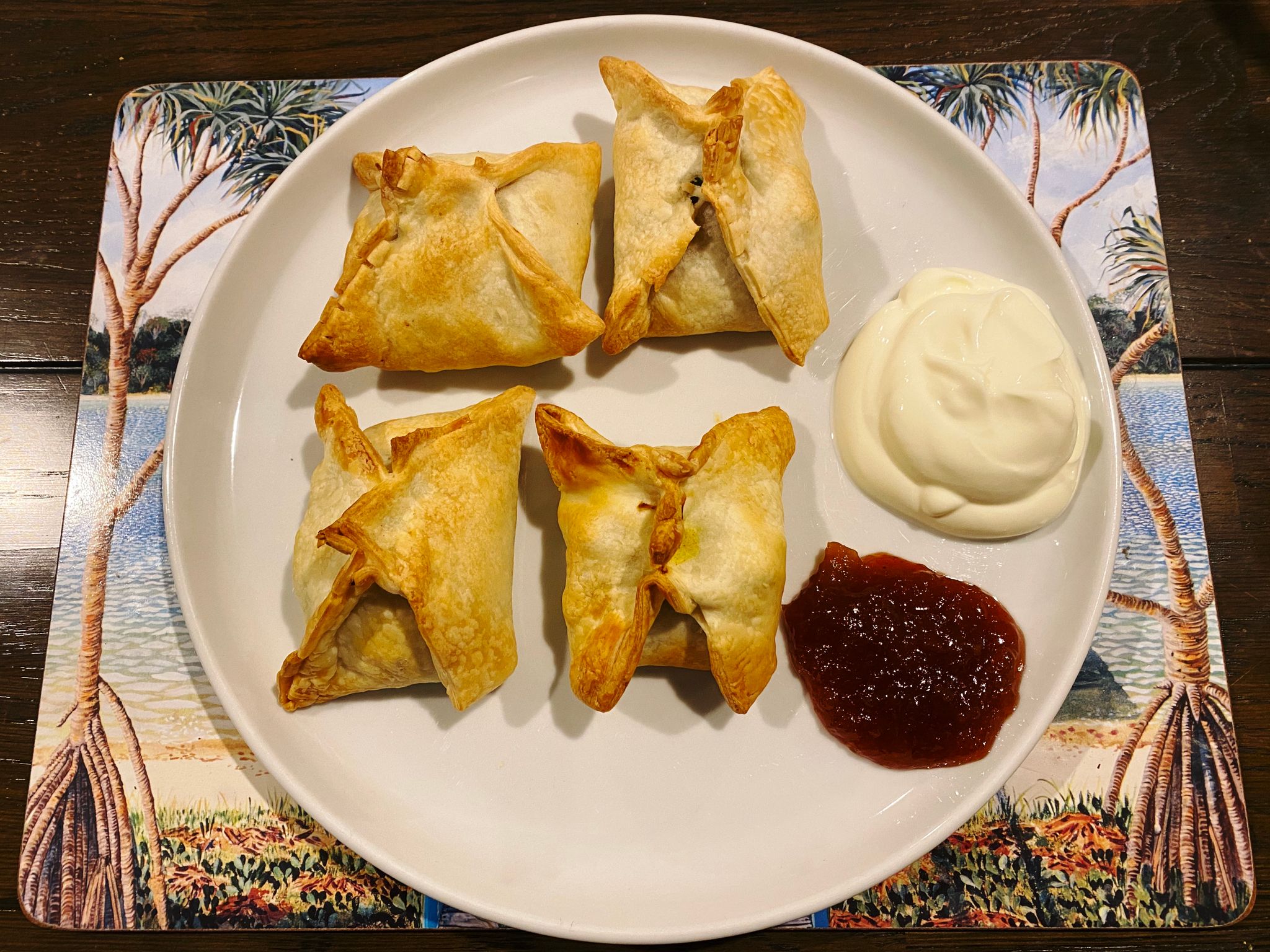 A photo of a plate with four (pyramid-shaped rather than triangular) samosas with a big dollop of yoghurt and another big dollop of mango chutney next to them.