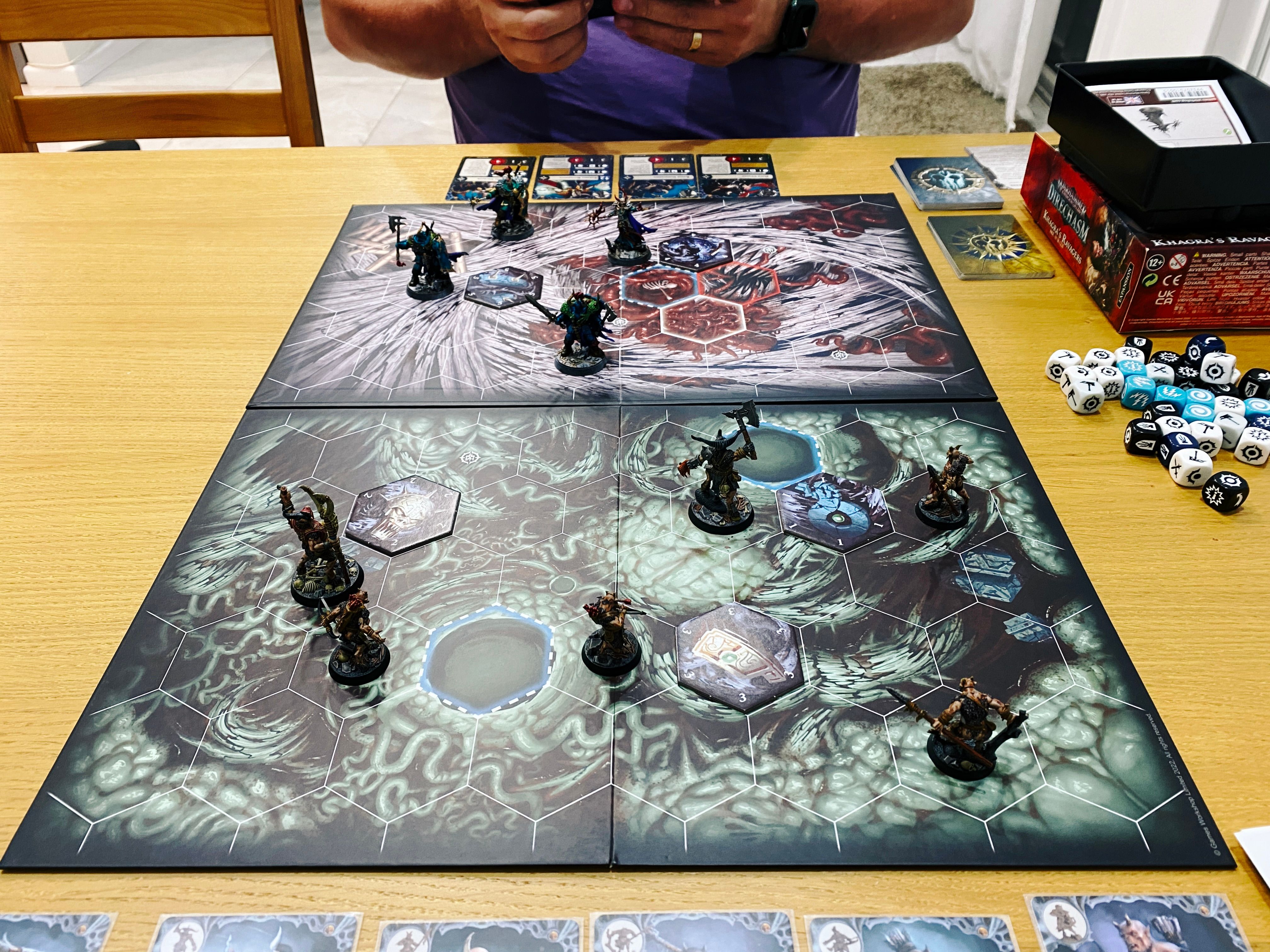 A photo of the game board of a Warhammer Underworlds game. The artwork on it looks like a bunch of gnarled roots and it's covered in hexagons that indicate distances. Closest to the camera are Grashrak's Despoilers, six beastmen wielding very crude-looking weapons and wearing not a huge amount of armour. Facing them are Khagra's Ravagers, four very heavily-armed- and -armoured human warriors.