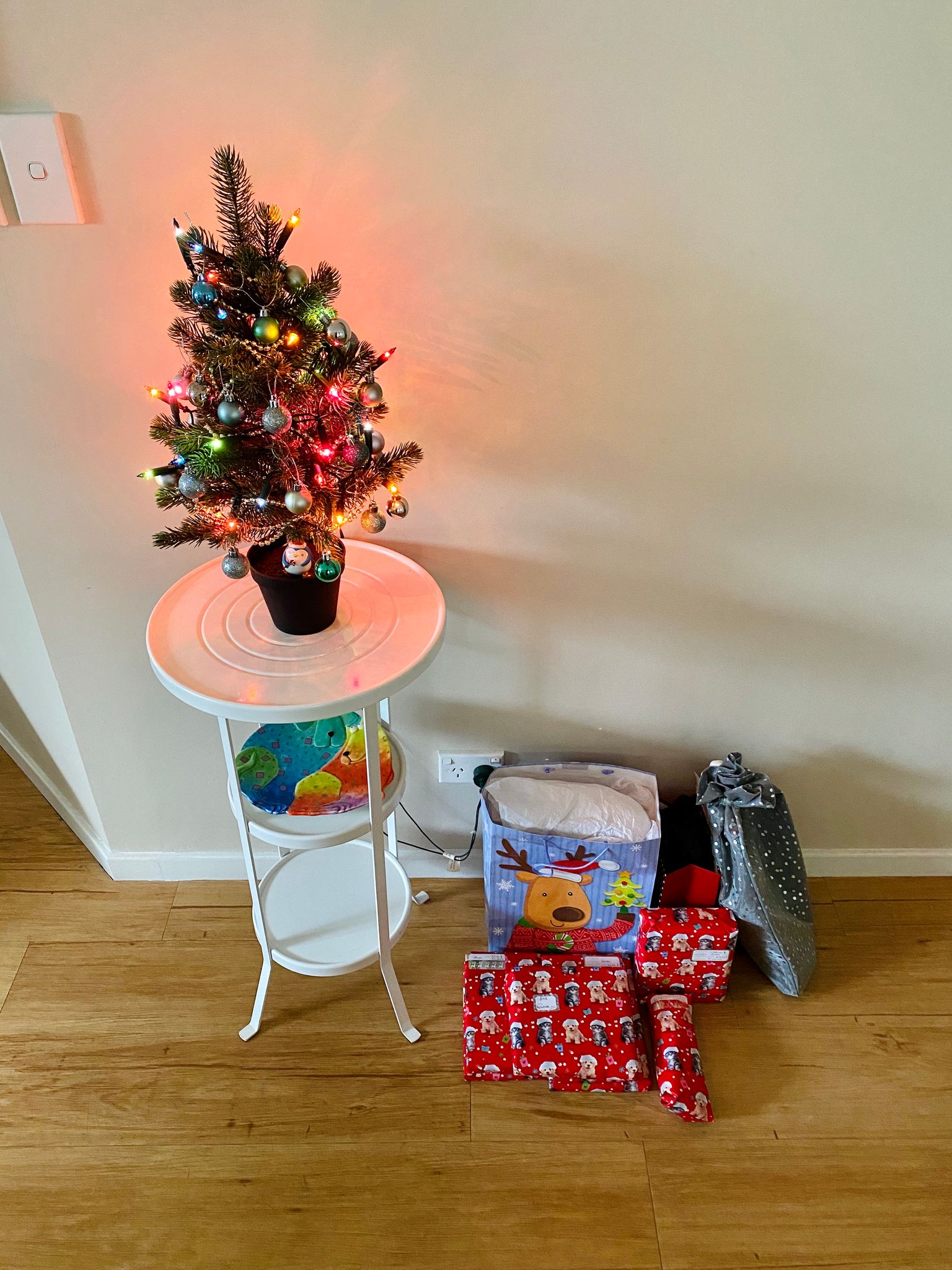 A small pile of Christmas presents sitting at the base of a round white pedestal, on top of which sits a very little (fake, plastic) Christmas tree covered in lights and baubles.