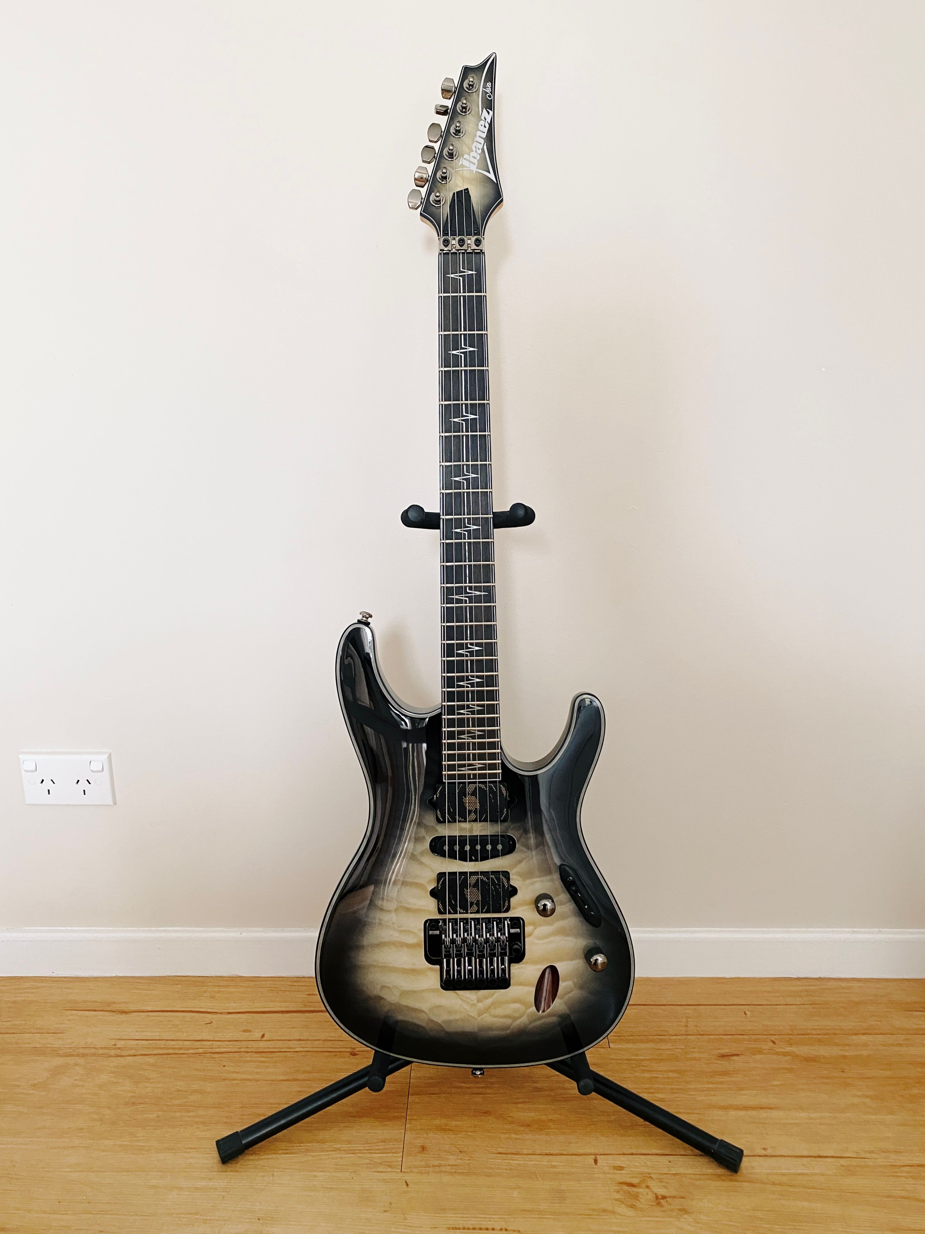 A photo of Ibanez's Nita Strauss signature "JIVA" electric guitar sitting on a guitar stand. The edges of the body are black and fade into the native maple in the middle, and down the fretboard where the fret markers are there's lines that look like an ECG readout.