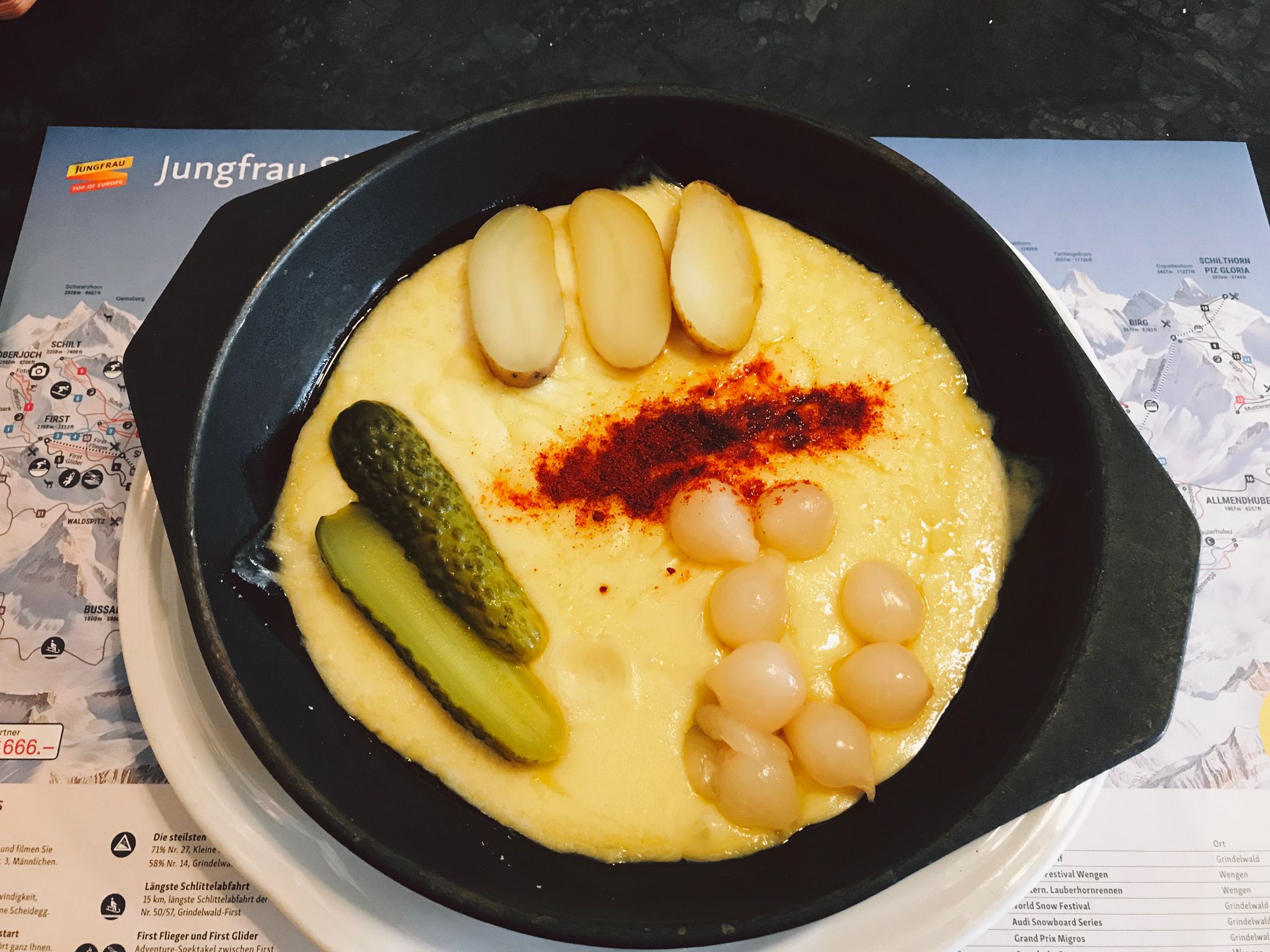 A photo of a cast-iron dish with the bottom covered in yellow raclette cheese, with potatoes, cocktail onions, and pickles on top.