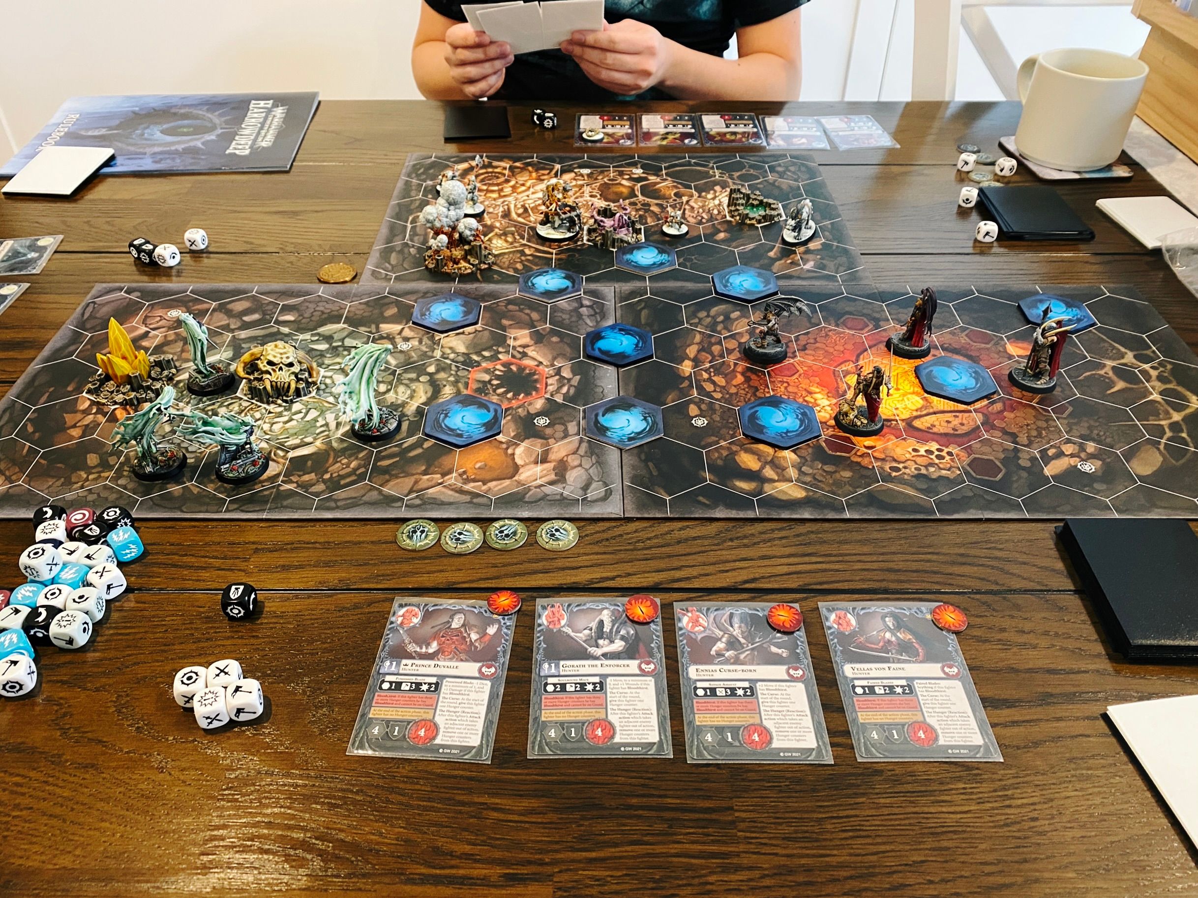 A photo of a three-player game of Warhammer Underworlds, a hex-based board game with miniatures and cards. Lady Harrow's Mournflight (four flowing spirits) are at the left, The Crimson Court (four elegant vampires in silver and bronze armour with red cloaks) are at the right, and across the other side of the board are Hrothgorn's Mantrappers (a huge ogre with a crossbow/bear trap launcher, a sabretooth tiger, and four goblins).