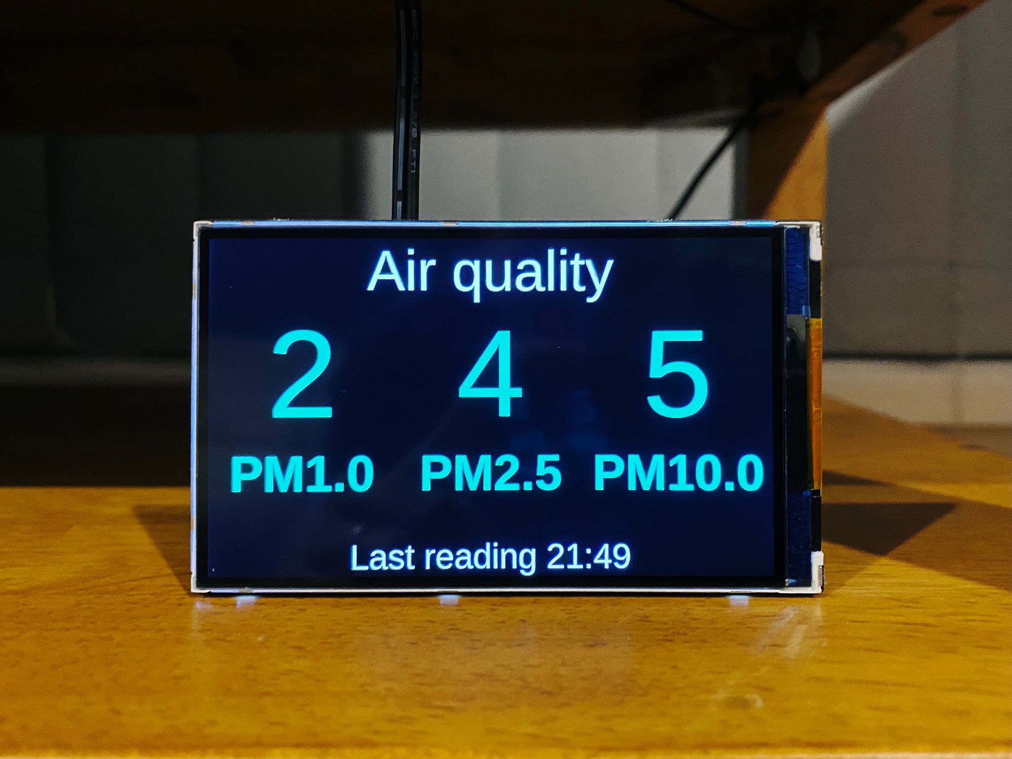 A photo of a 4" LCD display sitting on a shelf, showing PM1.0, PM2.5, and PM10 values.