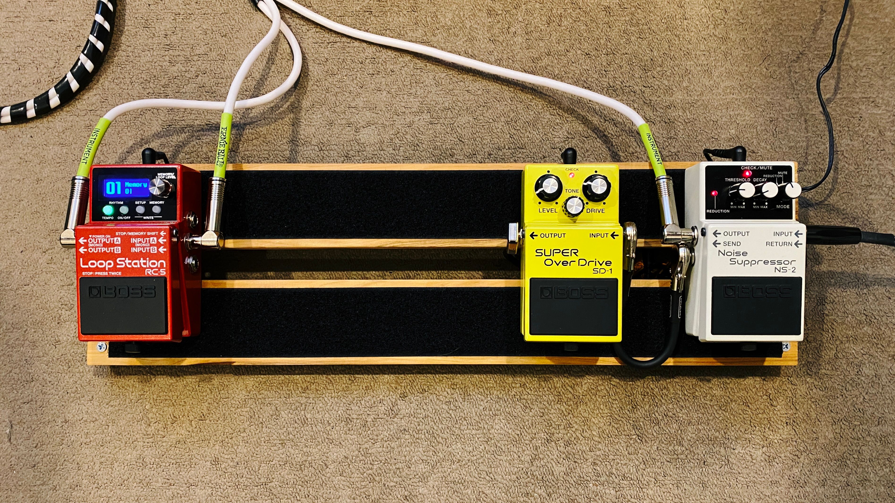 A photo of a DIY guitar pedalboard. It consists of two horizontal planks of wood screwed into two shallowly-angled pieces on either end. The two horizontal pieces have strips of velcro on them, and there's three pedals attached to it. The cables are extremely neat and tidy and mostly tucked away underneath the body of the board.