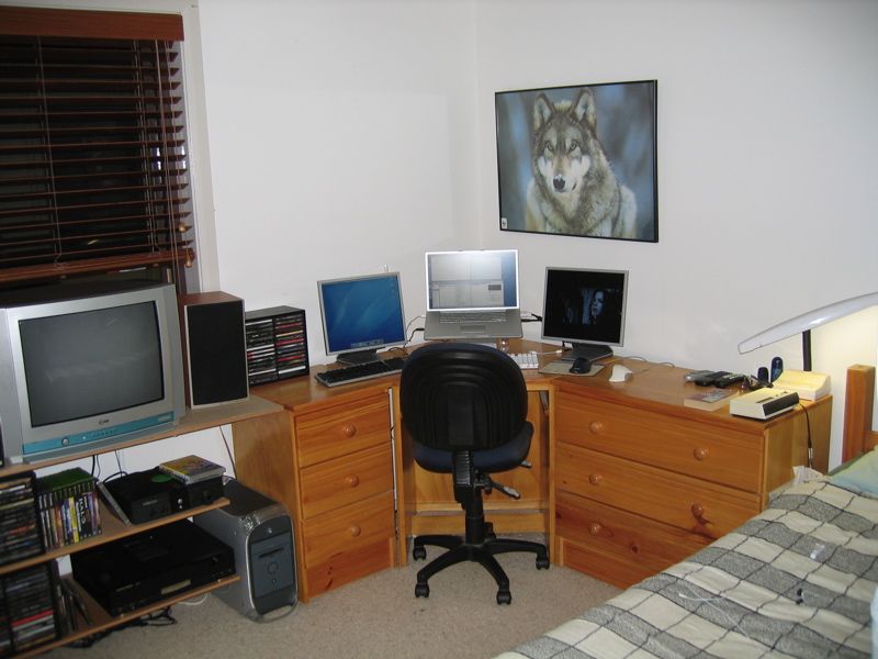 A photo of my room. There's a corner desk with a 17" PowerBook G4 flanked by two 17" 4:3 monitors, a Power Mac G4 on the floor next to the desk, a CRT TV on a set of wall-mounted shelves to the left of the desk, along with a speaker and an Xbox on the shelves underneath it. Also visible is a speaker next to the TV, and a CD rack full of CDs on the shelves under the TV.