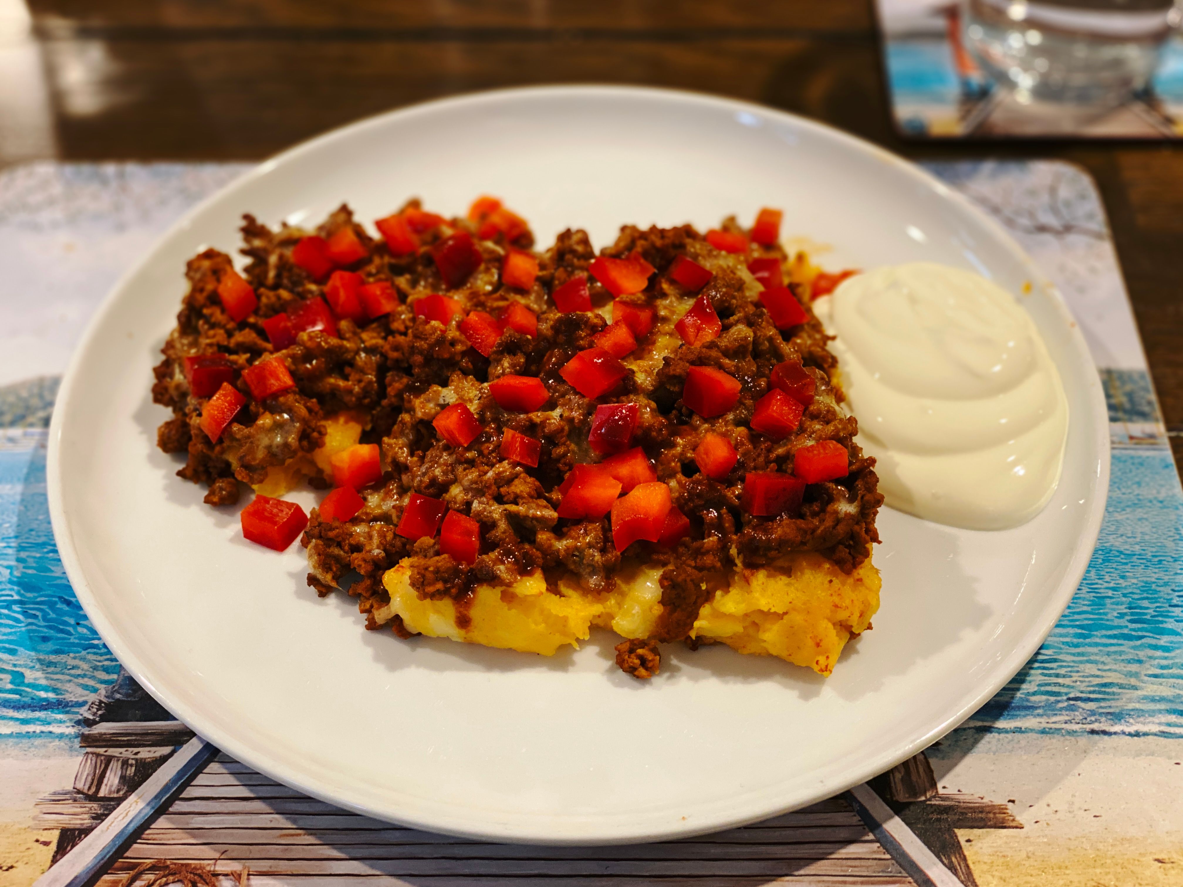 A photo of a dinner plate with a bed of golden mashed potato with a layer of beef mince on top, covered in chopped-up red capsicum and with a healthy dose of chipotle Tabasco sauce on top, and a big dollop of sour cream next to the mash.