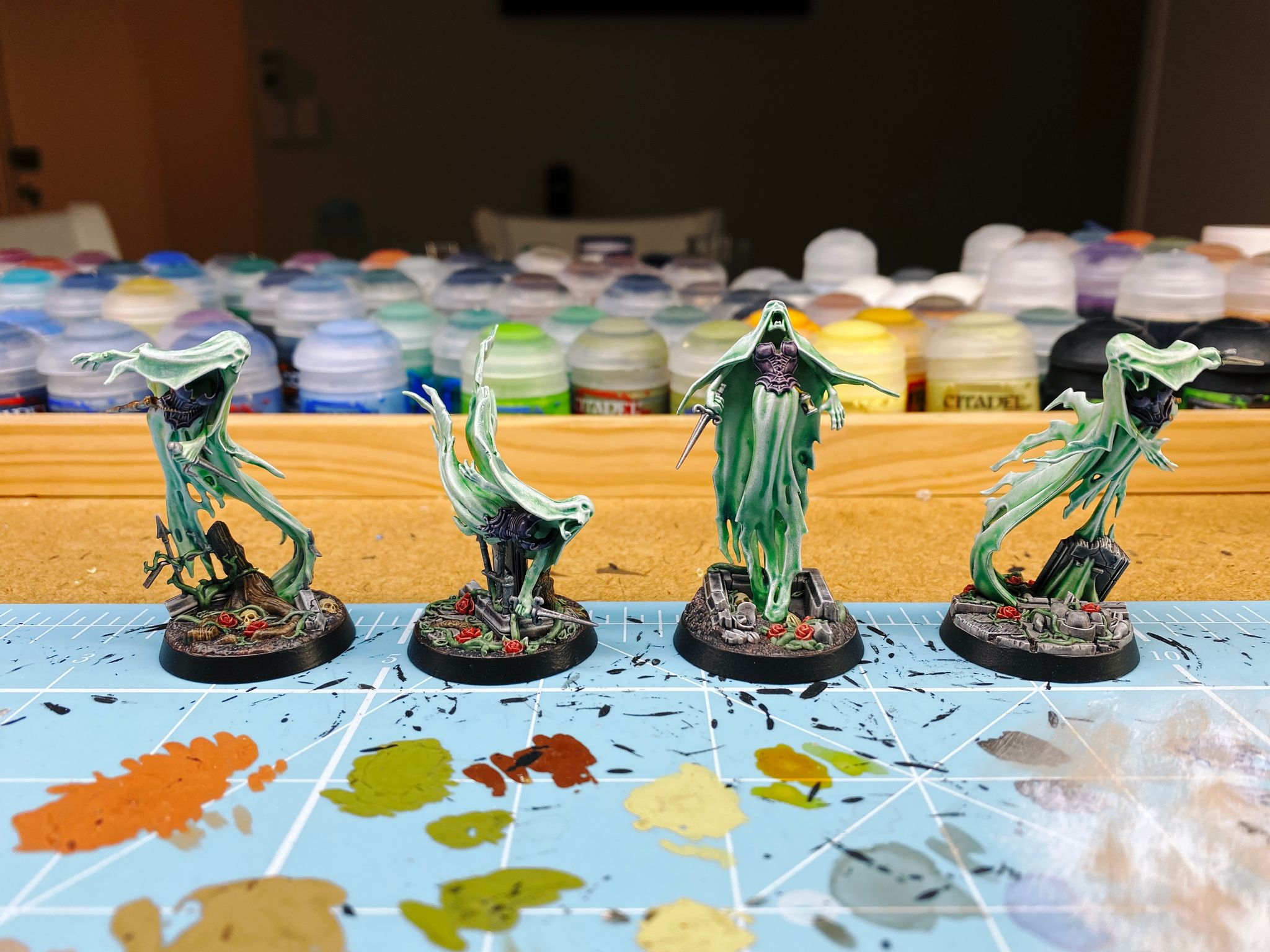 A photo of the four miniatures of Lady Harrow's Mournflight from Warhammer Underworlds. They're spirits and modelled to look like they're very ephemeral and wispy and flowing. The wispy bits are a sickly green colour with a smooth gradient into white at the edges and raised areas, they have deep purple corsets, and the bases look like a ruined graveyard.