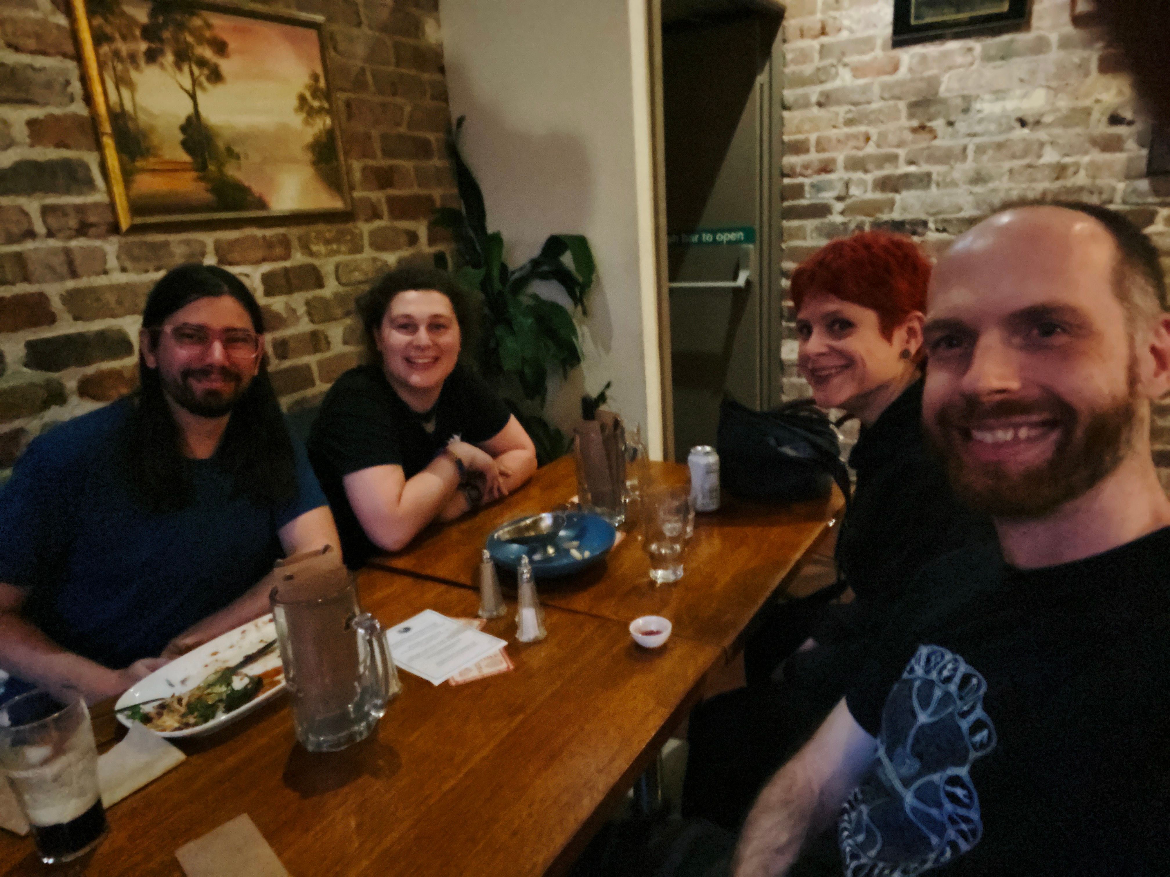 A selfie of four people sitting around a restaurant table smiling at the camera. Shlee has long dark hair and a beard, s0 has long curly hair pulled back into a bun, yayKM has short bright red hair, and I have short brown hair and a red beard.