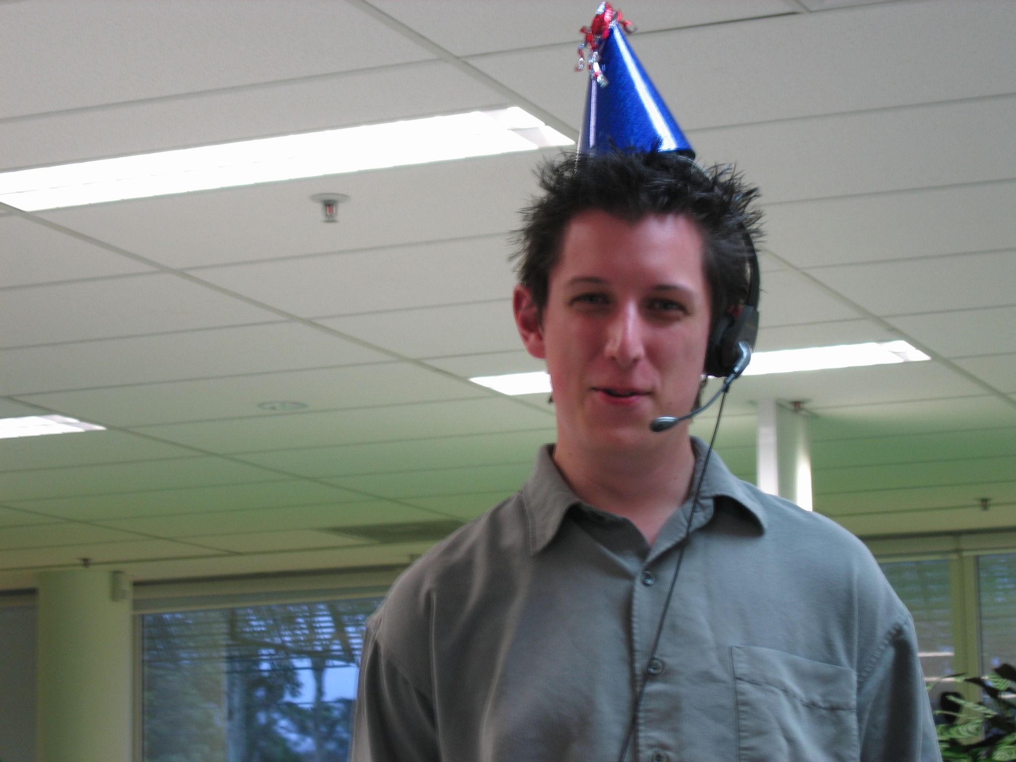 A photo of a clean-shaven white man in a khaki-coloured collared shirt wearing a call centre headset and a conical party hat.