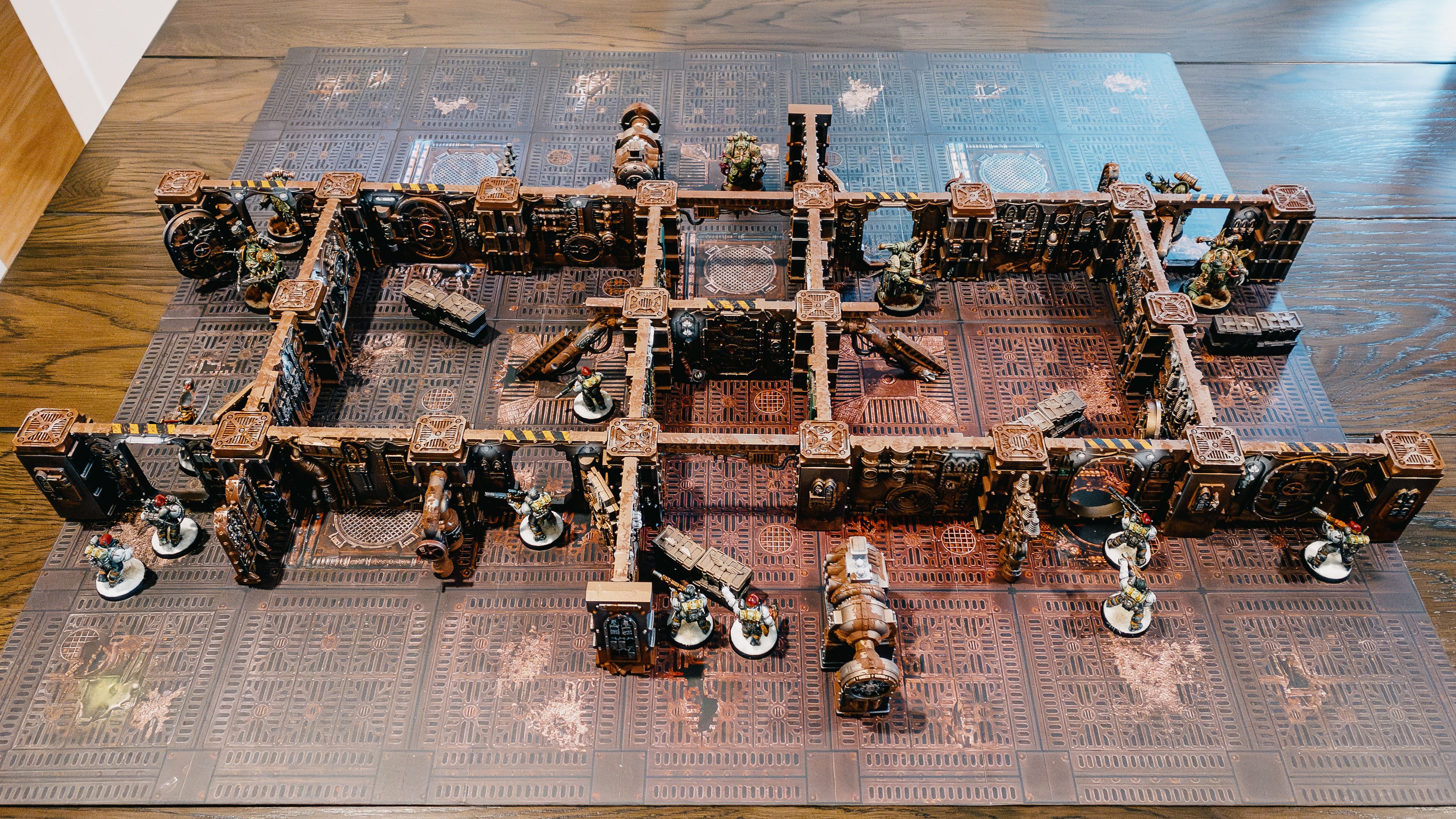 A photo of a bunch of steampunk-looking terrain on a game board, it's all walls and put together to look like the interior of a gothic-slash-steampunk starship. The terrain is all rusty-looking like it's been properly abandoned for centuries. Closest to the camera are the warriors of the Astra Militarium, ten heavily-armoured women in white-coloured armour wielding various guns. On the far side are six Plague Marines, huge superhuman warriors in green armour that have all sorts of gross mutations.