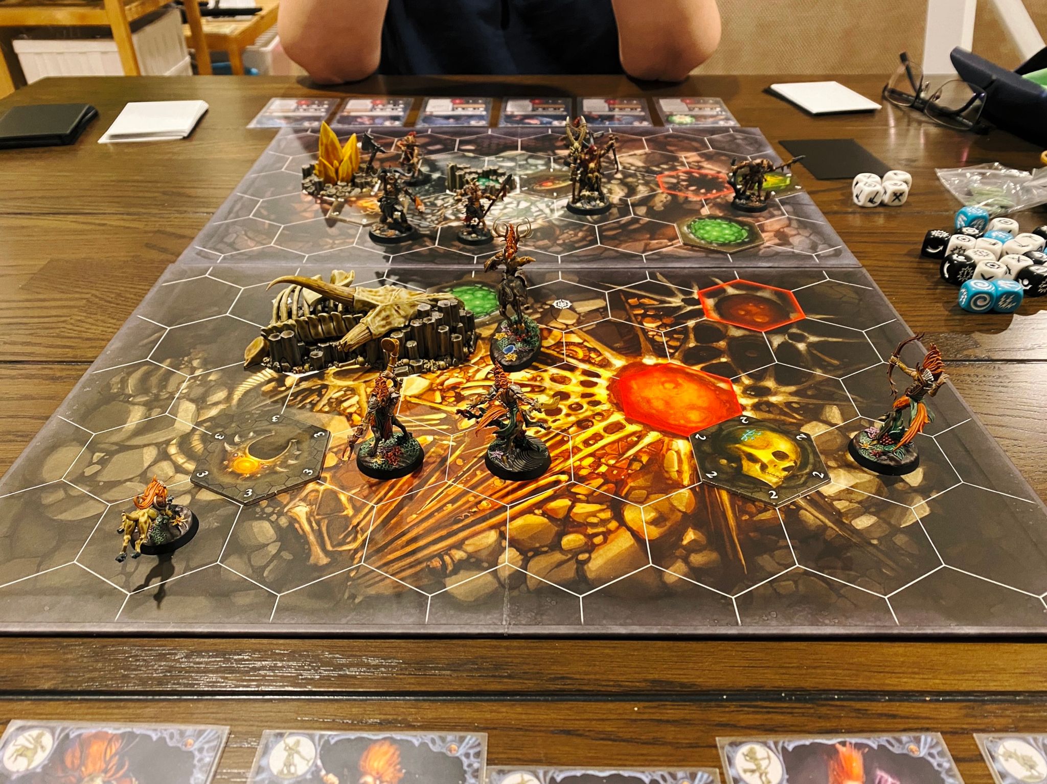 A photo of a Warhammer Underworlds game board, it has hexagonal tiles on it and the artwork on the board looks like the inside of a cave. Skaeth's WIld Hunt are on one side, and they're all elegant and flowing wood elf-like creatures, with the leader being a centaur, and on the other side Grashrak's Despoilers are ugly and brutish-looking goat-men.