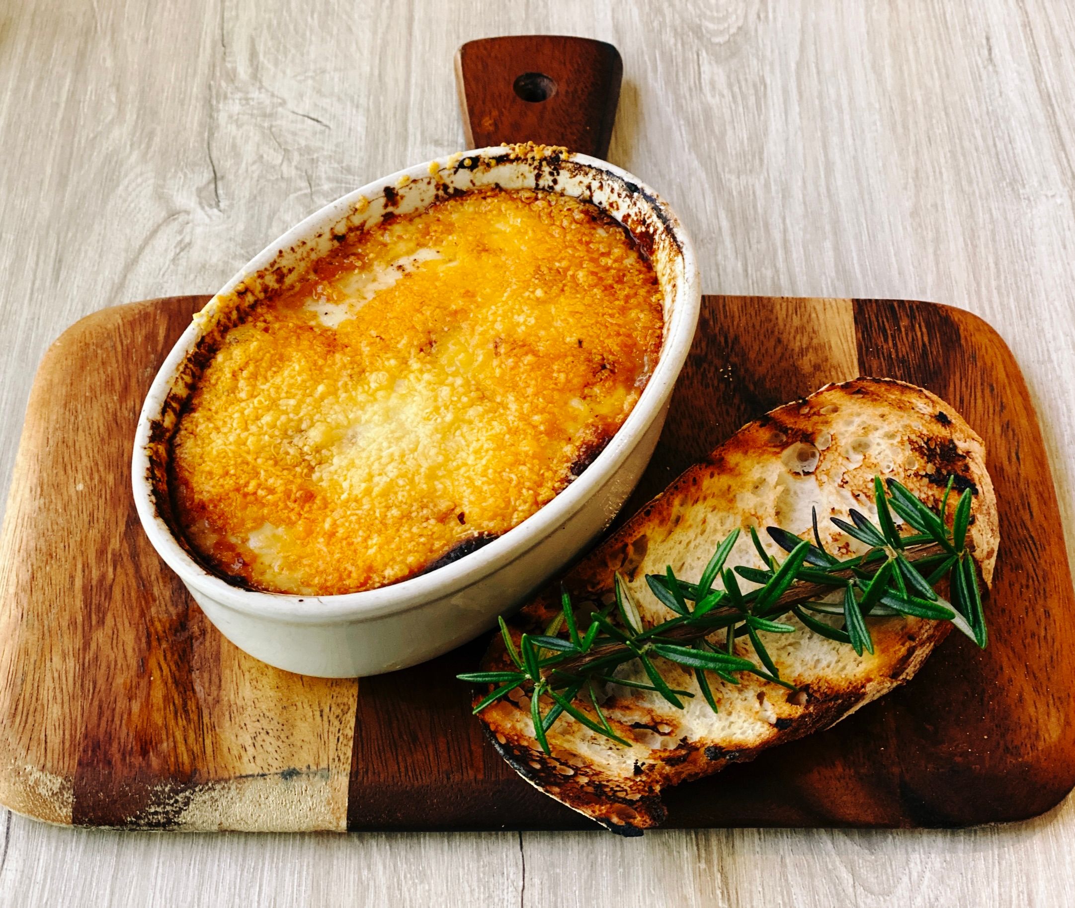 A photo of a deliciously golden brown moussaka in an oval dish alongside a piece of toasted bread.