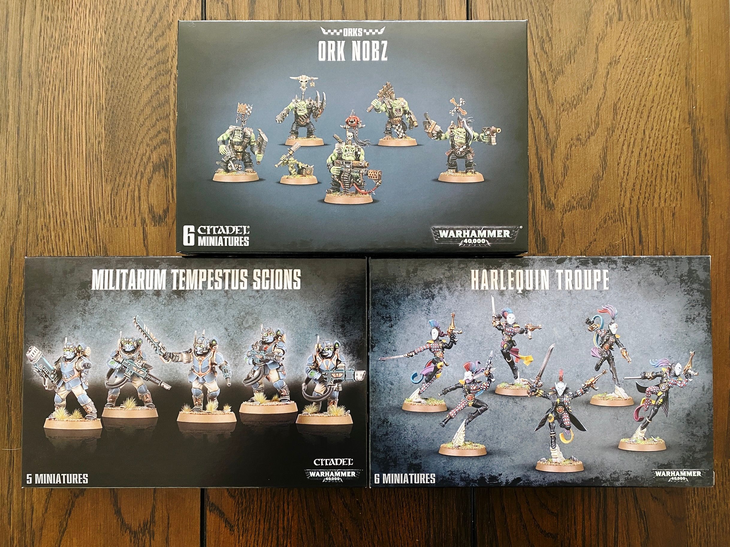 A photo of three boxes of Warhammer 40,000 miniatures: Ork Nobz (thuggish-looking greenskinned aliens with big teeth and a motly assortment of armour and guns), Astra Militarum Tempesus Scions (armoured human soldiers) and a troope of Aeldari Harlequins (lithe humanoid aliens in multicoloured clothing all in dynamic poses, all wearing white harlequin masks).