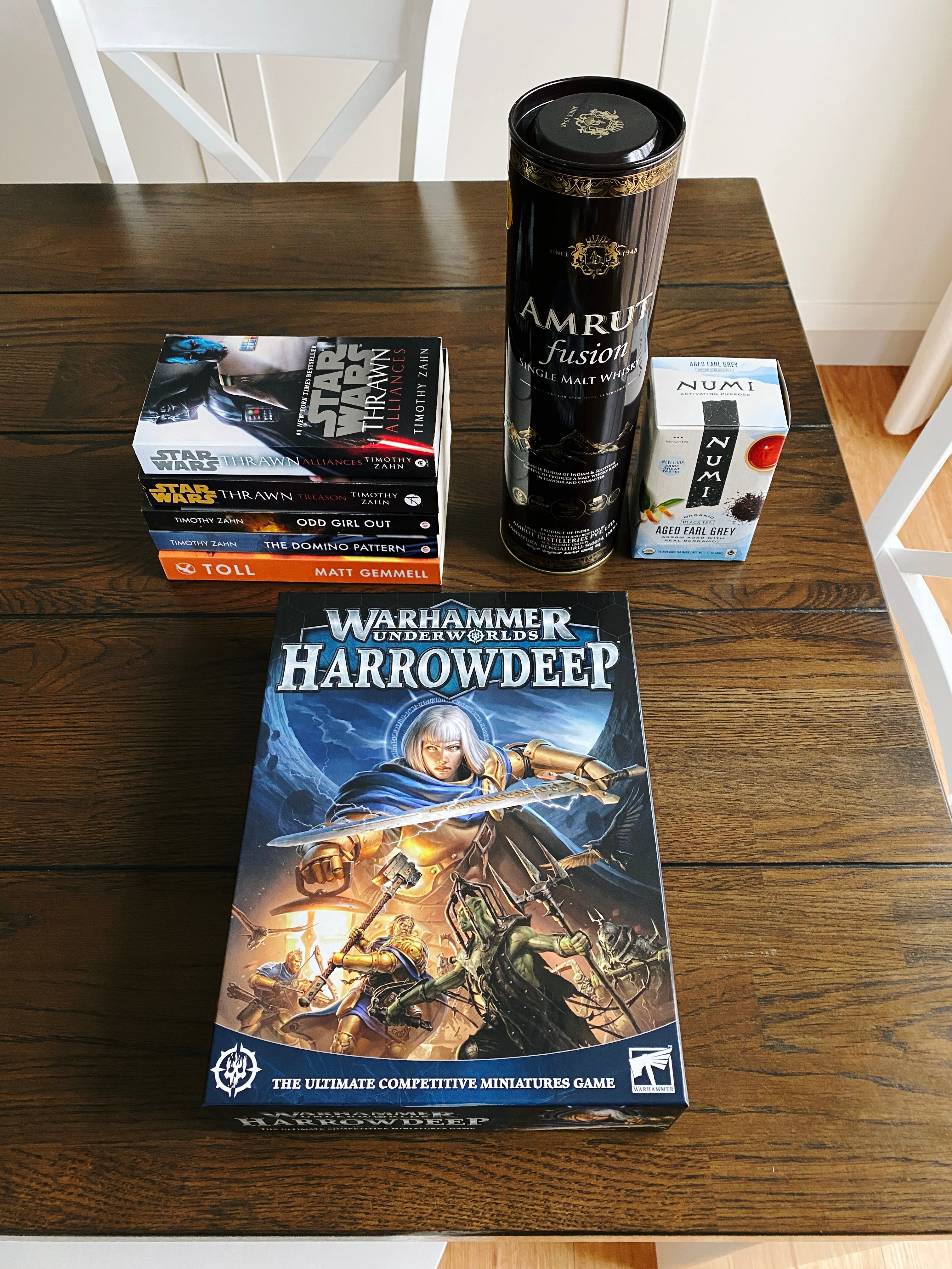 A photo of five novels, the latest edition of Games Workshop's "Warhammer Underworlds" game called "Harrowdeep", a cylindrical container with a bottle of whisky in it, and a box of "aged" Earl Grey tea.