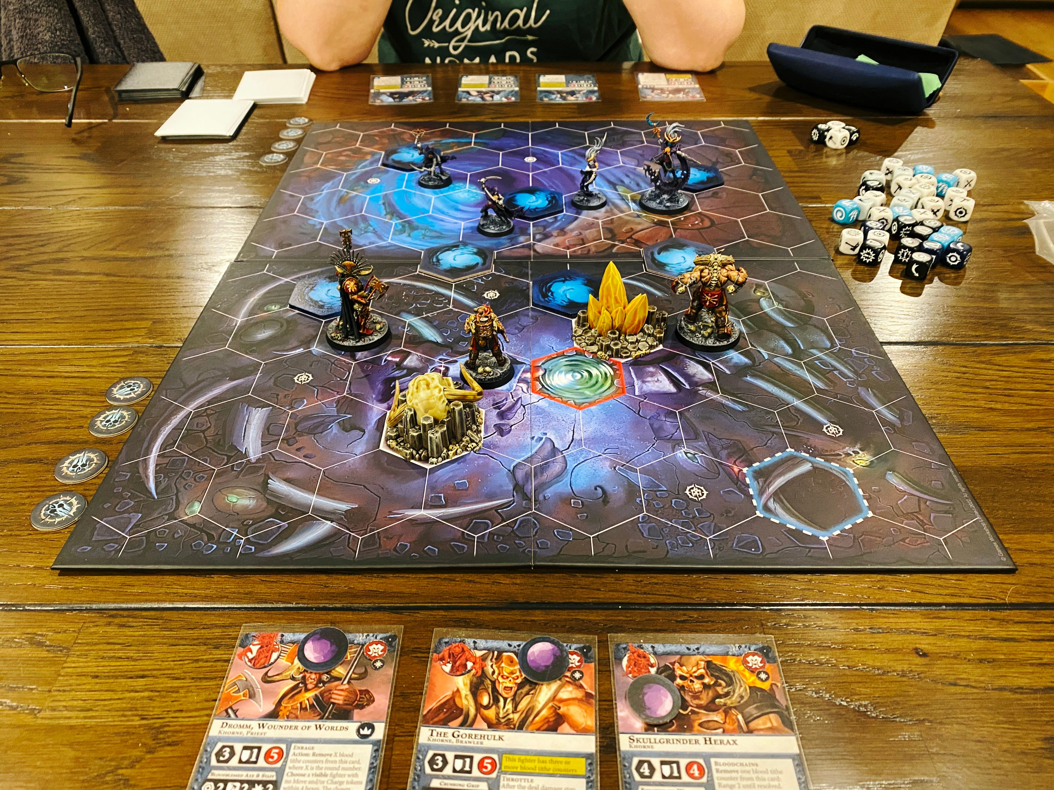 A photo of a game of Warhammer Underworlds. It's a miniatures game played on a board with hex tiles on it, on my side are three very large armoured warriors, and facing them are four lithe and much more lightly armoured elves.