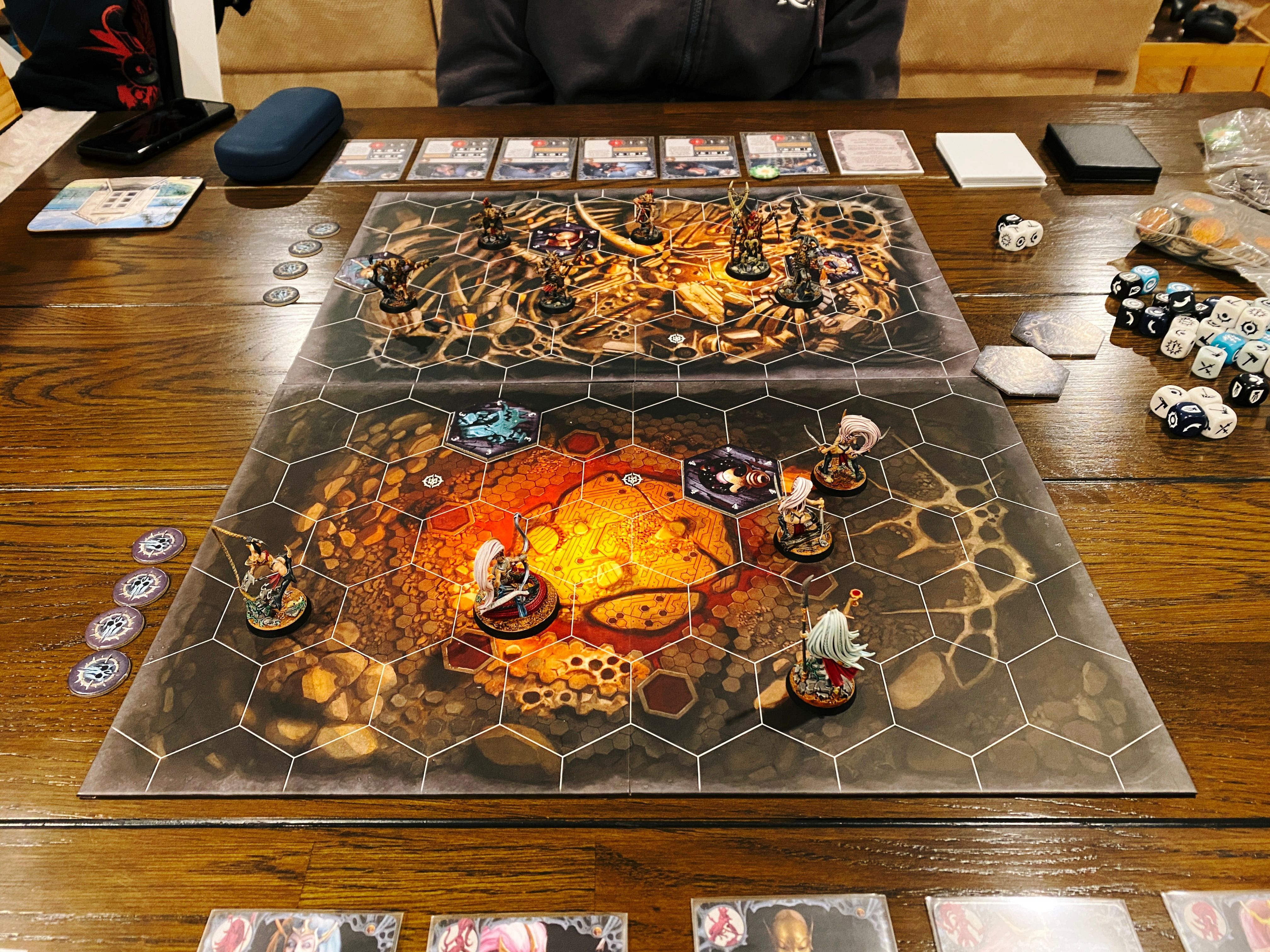 A photo of a Warhammer Underworlds game board. It has hex tiles on it, and the artwork looks like the inside of a cave. Closest to the camera are Morgwaeth's Blade-coven, a five-fighter warband consisting of lithe female elves who aren't wearing a whole lot of armour. At the other end of the board are the six goatmen of Grashrak's Despoilers: two are larger and more intimidating looking, one is holding a giant axe in the air and is gripping the head of someone he just decapitated, and the other four are smaller and shabbier, wielding bows and spears.