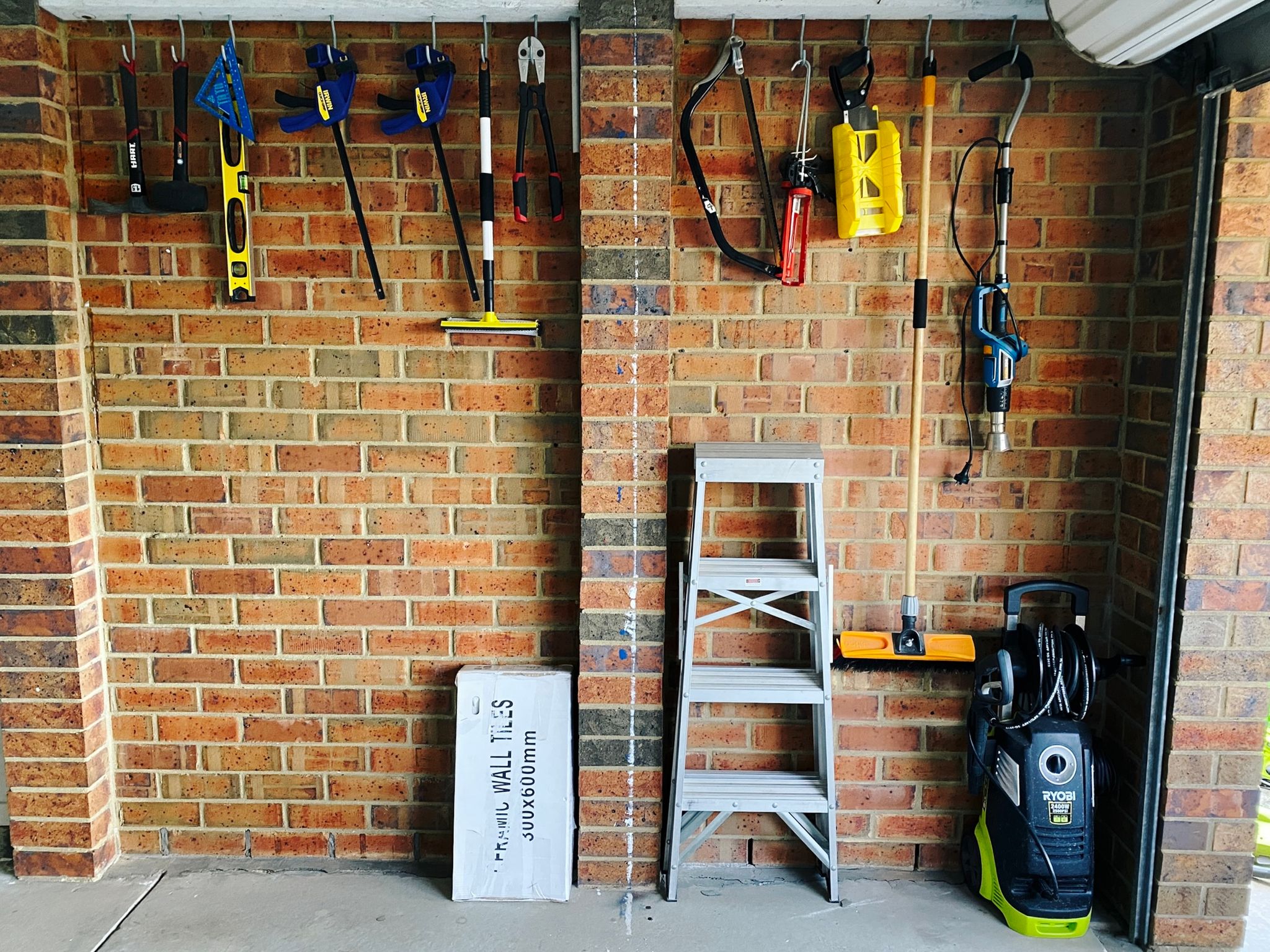 A photo of the inside side wall of our garage with a bunch of vertically-hanging hooks attached to two wooden planks up near the ceiling. There's various tools hanging off them (a couple of saws, a couple of clamps, a level, bolt cutters, a broom, etc.).