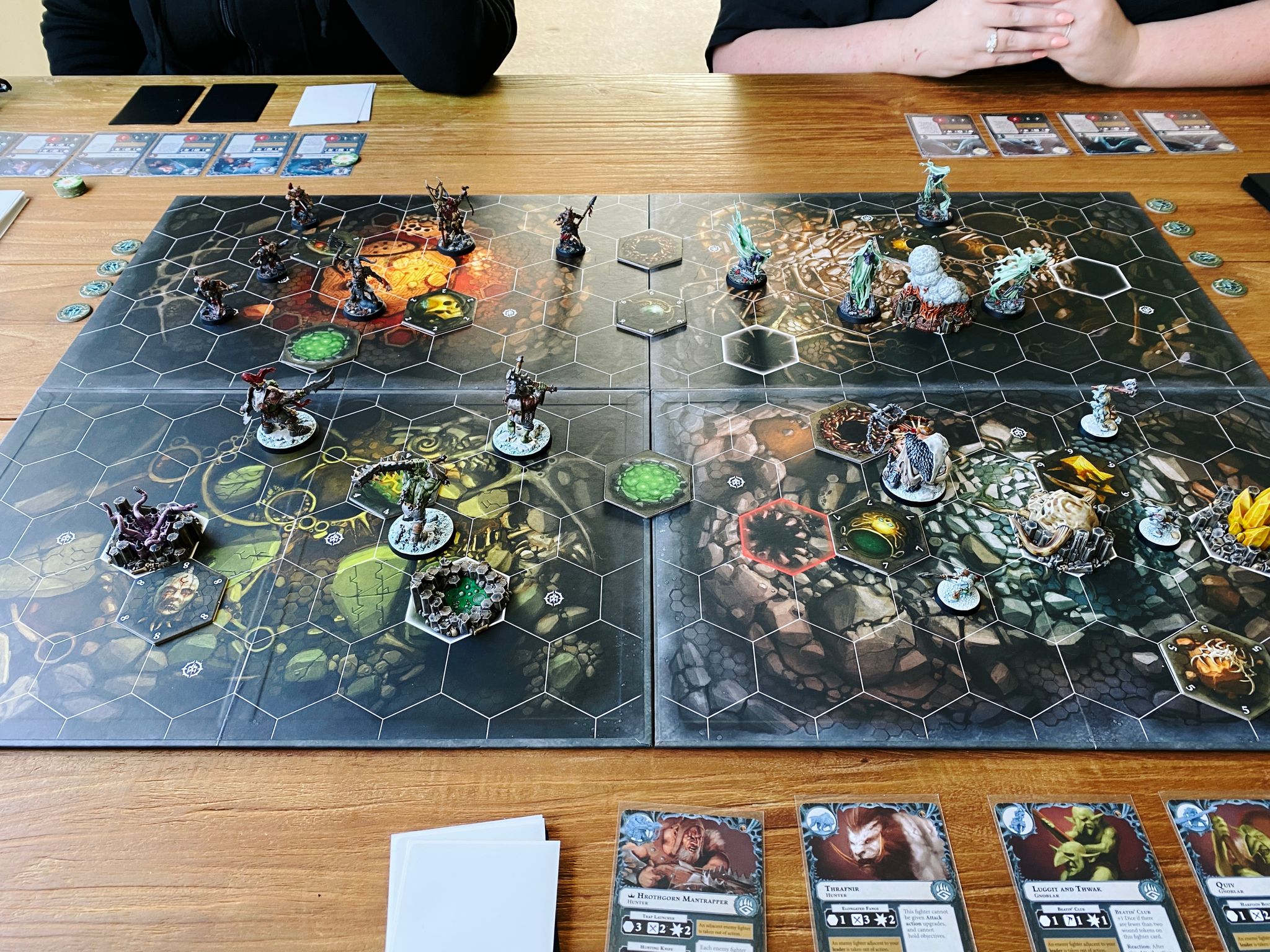 A photo of a four-player game of Warhammer Underworlds. The board has hexes marked out on it, at the bottom right are Hrothgorn's Mantrappers (a big ogre and 4 goblins), bottom left at Morgok's Krushas (3 heavily-armoured orks), top-left are Grashrak's Despoilers (6 goat people), and top-right is Lady Harrow's Mournflight (4 ghosts).