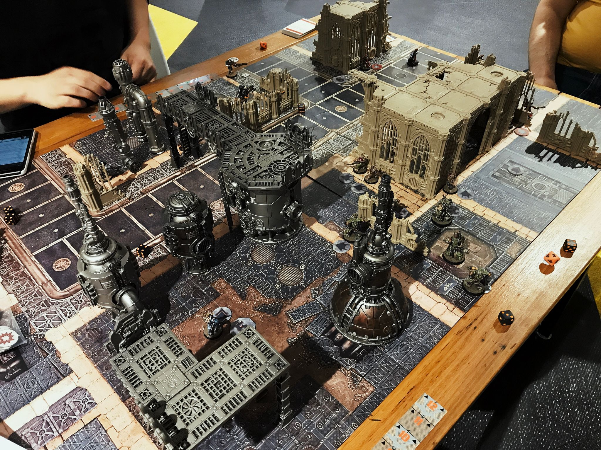 A photo of a Warhammer 40,000: Kill Team game. It's on two of the standard-size game boards, the left half has very industrial-looking terrain, all metallic and pipes, the right half's terrain is ruined gothic church-style buildings. A team of Death Guard at are the bottom, Necrons at the top, and Adeptus Mechanicus at the right.