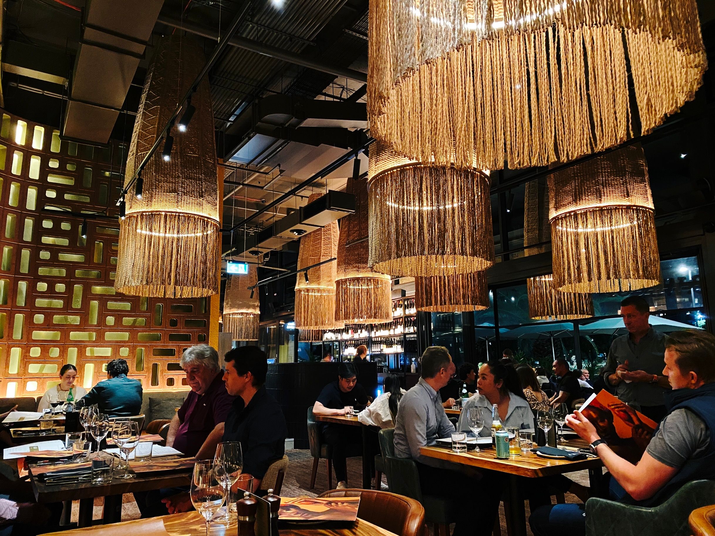 A photo of inside a restaurant with people sitting at ables. The light levels are fairly low, and there's BIG wicker light fixtures hanging down from the ceiling with tassels hanging from the bottom of the wicker. The wall at the back-left is made from concrete blocks with holes in the middle, and behind THOSE is another wall that's been lit up.
