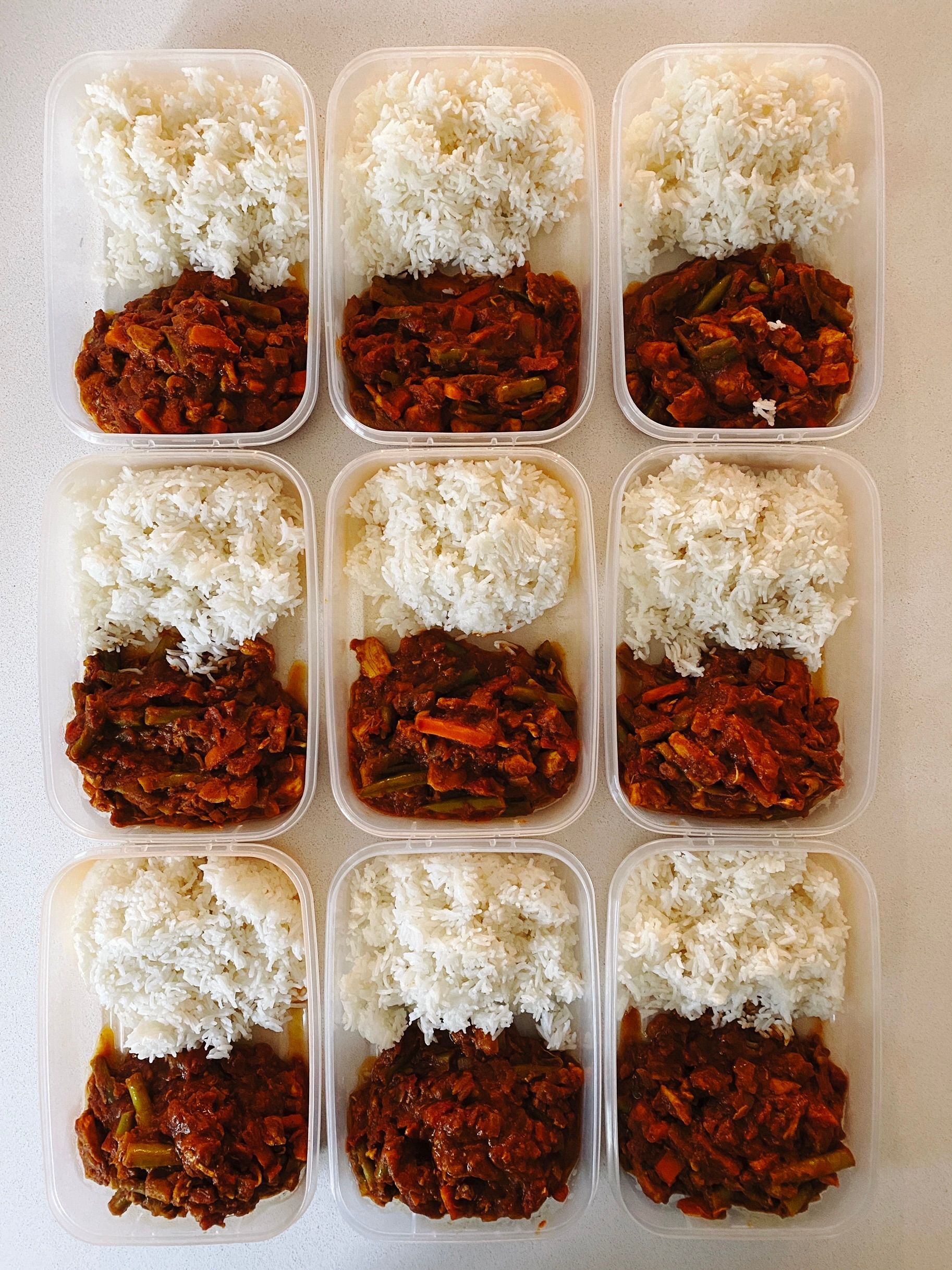 A photo of nine tupperware containers, each with half rice and half tomato-based chicken curry with beans and carrots on it.