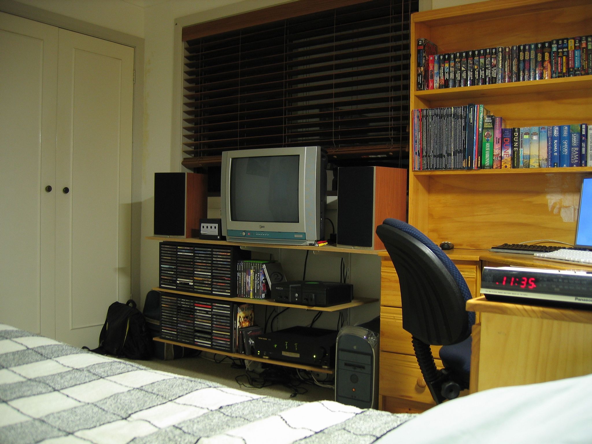 A photo taken with the camera sitting on a bed, looking towards the opposite wall of the room. There's a window with brown wooden venetian blinds, in front of the window are three shelves mounted to the wall. On the top shelf is a CRT TV, two bookshelf speakers, and a Nintendo GameCube. On the second shelf is a CD rack full of CDs, the jewel cases of a selection of Xbox and GameCube games, and an original Xbox. On the bottom shelf are more CDs, and a stereo amplifier. Next to the shelves are a graphite Power Mac G4, and next to that is a corner desk with tall bookshelves on top of the desk, fulled with books.