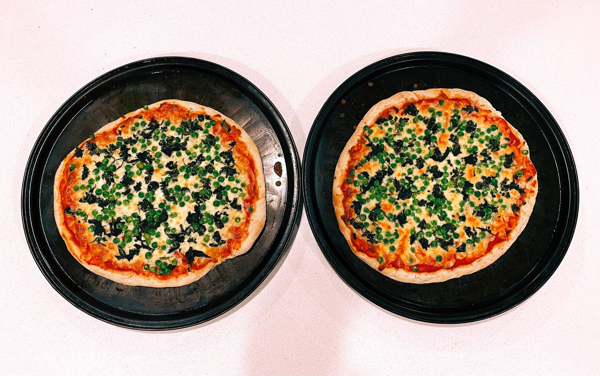 A photo of two pizzas with spinach and peas on them, sitting on pizza trays.