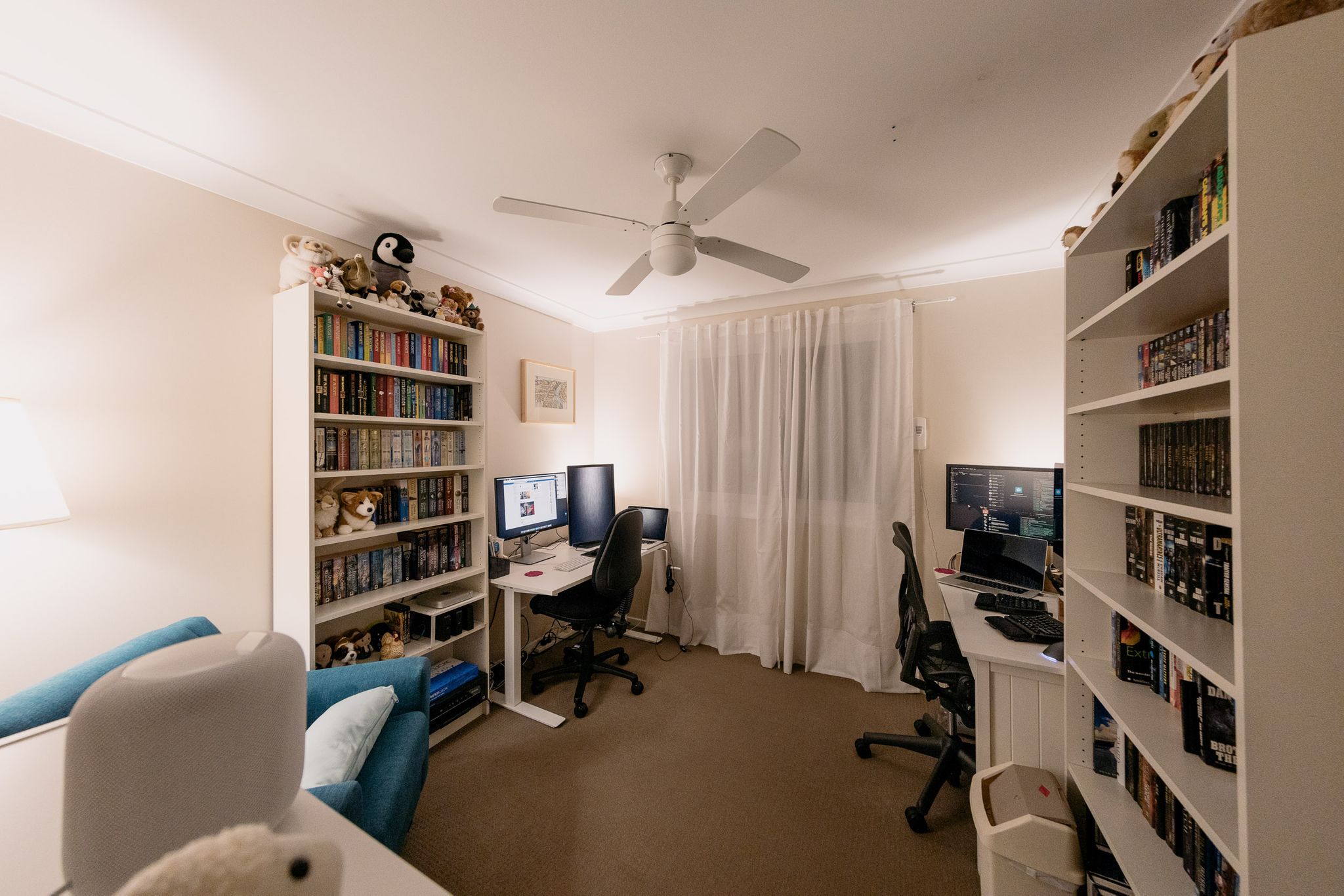 A wide-angle photo of our office. At the left and right walls are two white bookshelves with lots of books on them, right next to both are two desks with computers on them. There are stuffed animals on the top of the bookshelves, and a half-height bookshelf immediately to the bottom-left of the photo with a white Apple HomePod sitting on it.