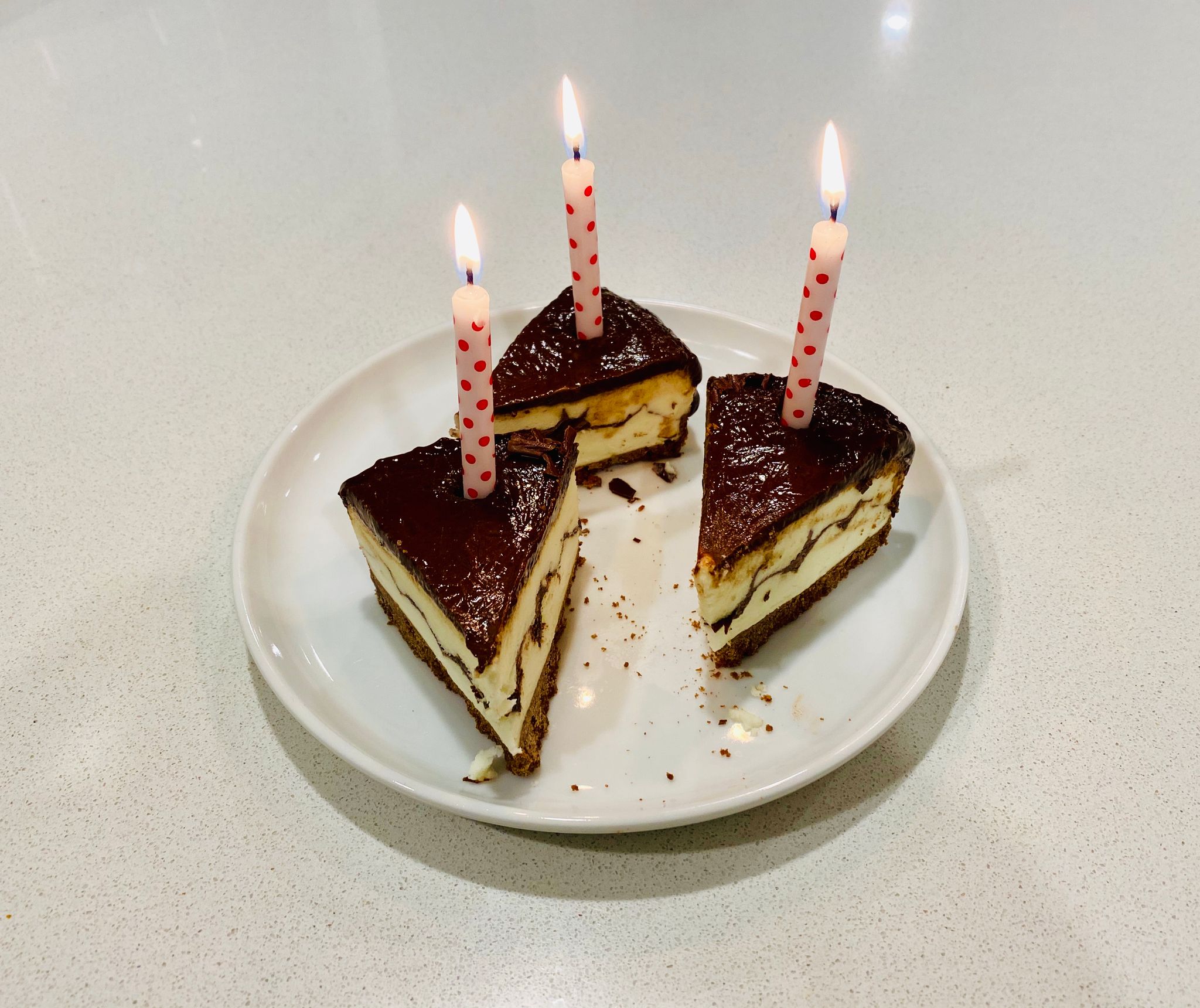 A photo of three small slices of chocolate-covered cheesecake with a lit candle on each piece.