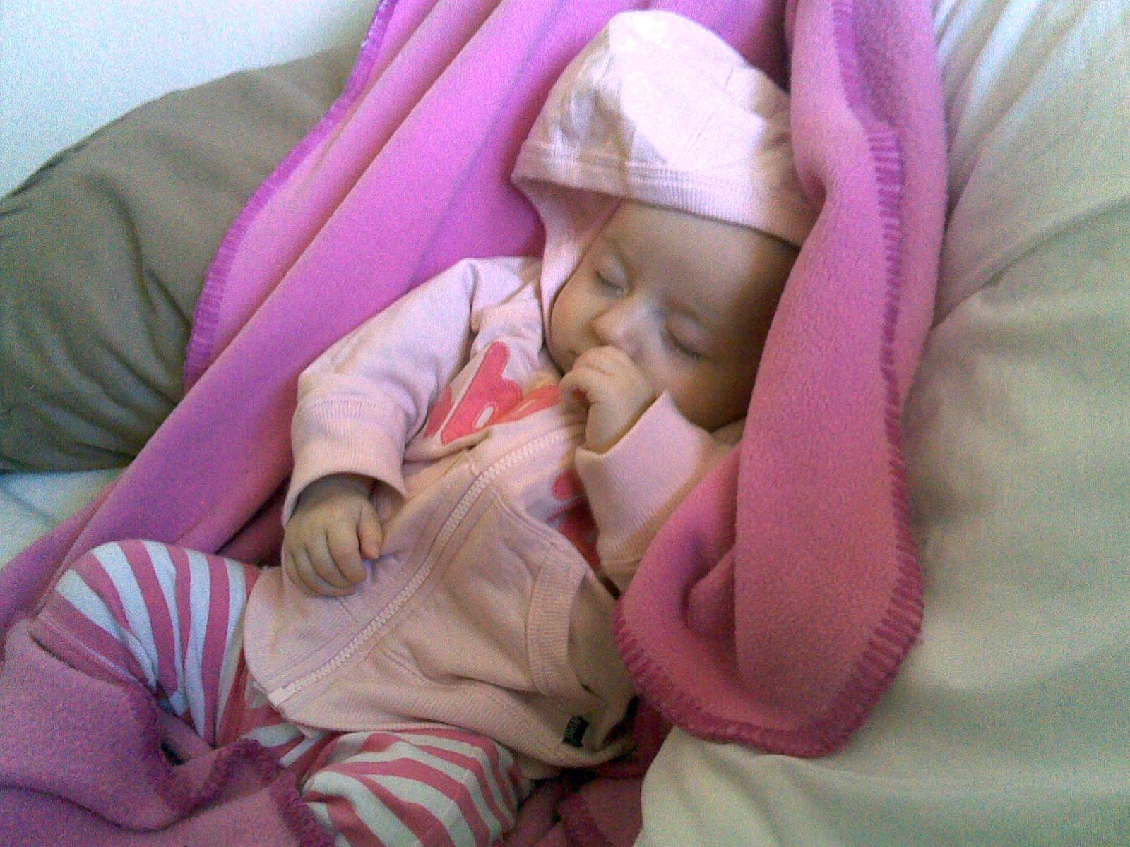 A photo of a small baby lying on a pink blanket with pink and white stripy pants on, and a pink hoodie. She's asleep and has her thumb in her mouth.